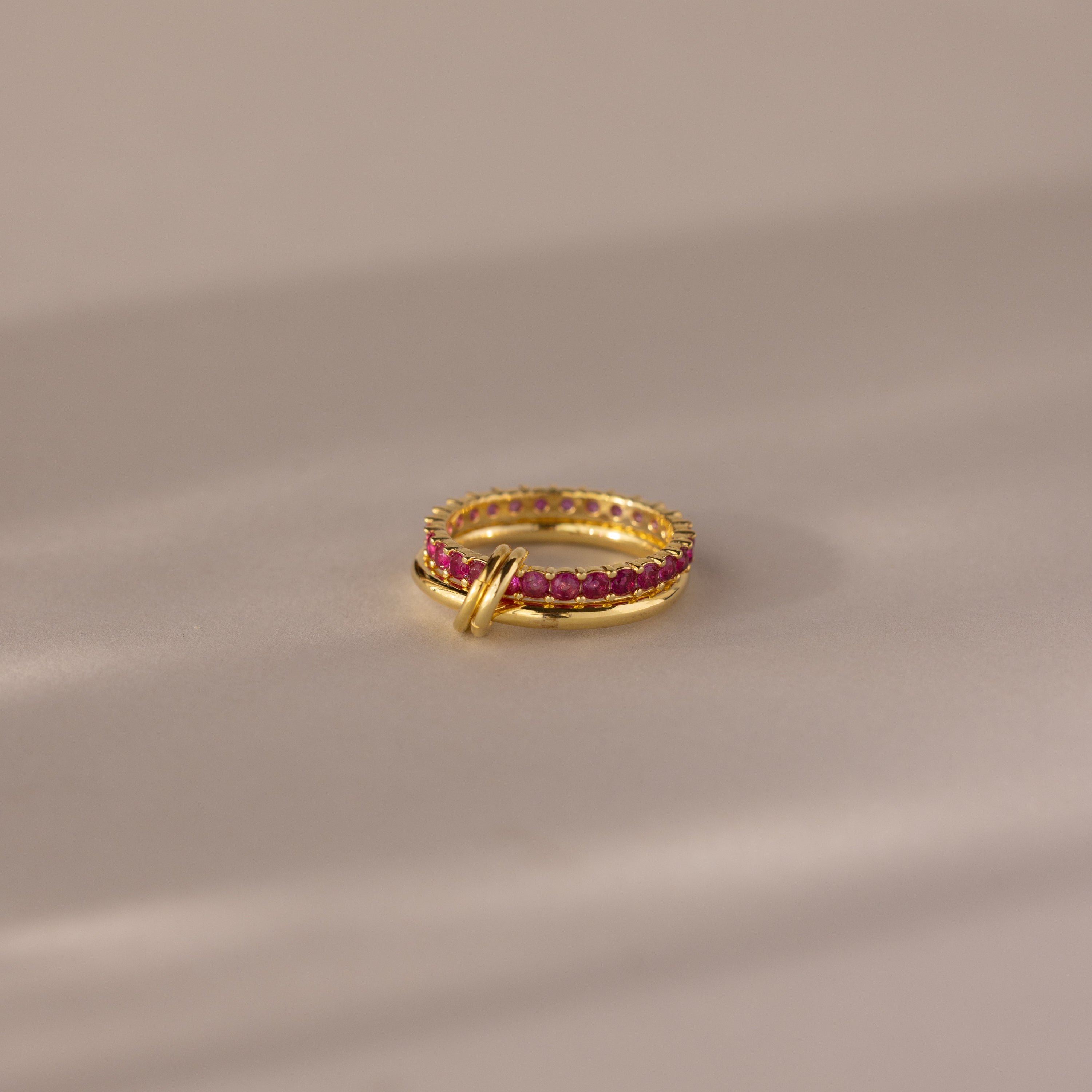 The Duo Birthstone Tennis Linked Ring Set in ruby (July) features minimalist gold rings with red gemstones, displayed on a soft beige surface under gentle lighting.