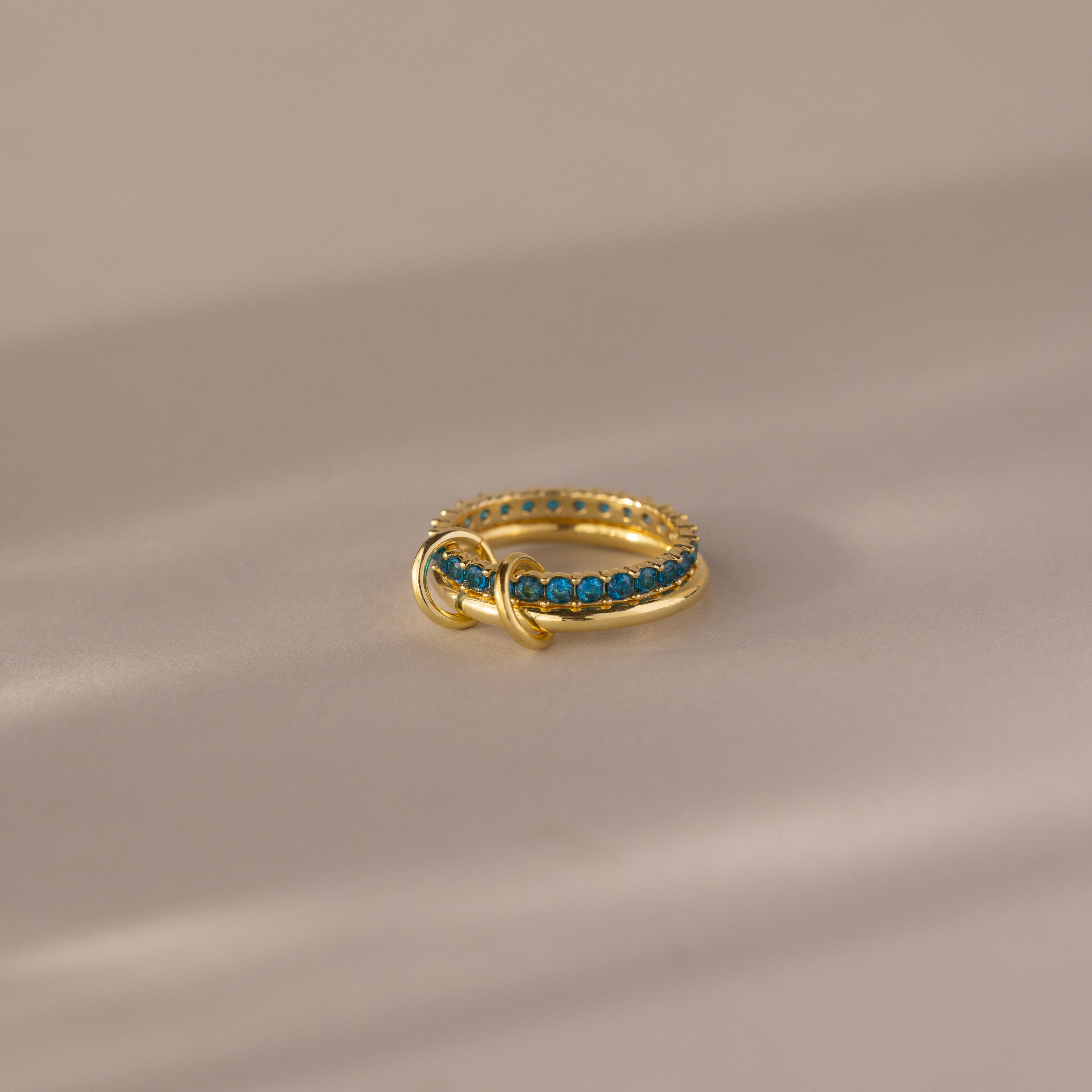 The Duo Birthstone Tennis Linked Ring Set in Blue Zircon (December) features blue gemstones set in a row on gold, displayed on a neutral, softly lit surface.