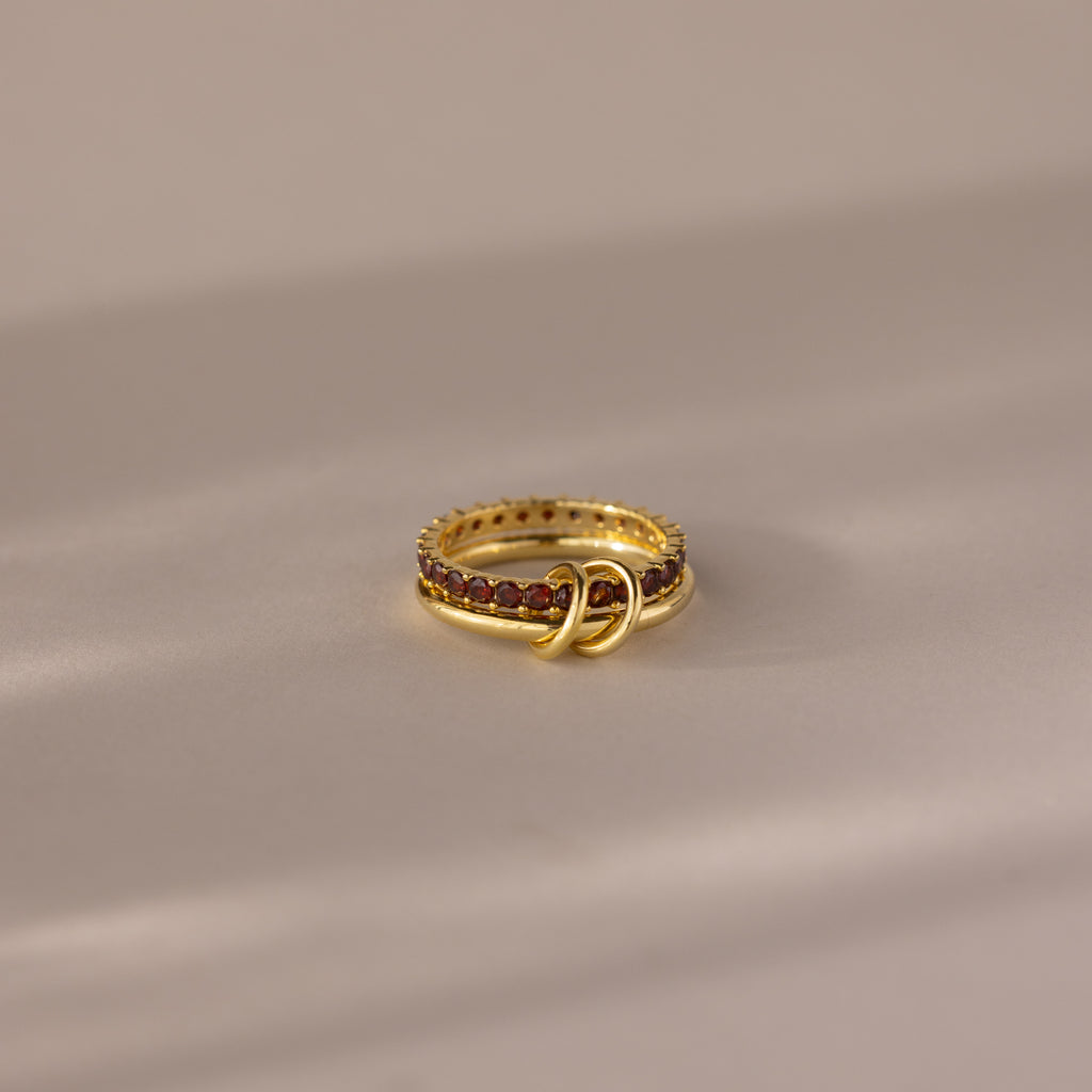 The Duo Birthstone Tennis Linked Ring Set in Garnet (January) features a minimalist, looped design with red gemstones, displayed on a soft neutral surface.