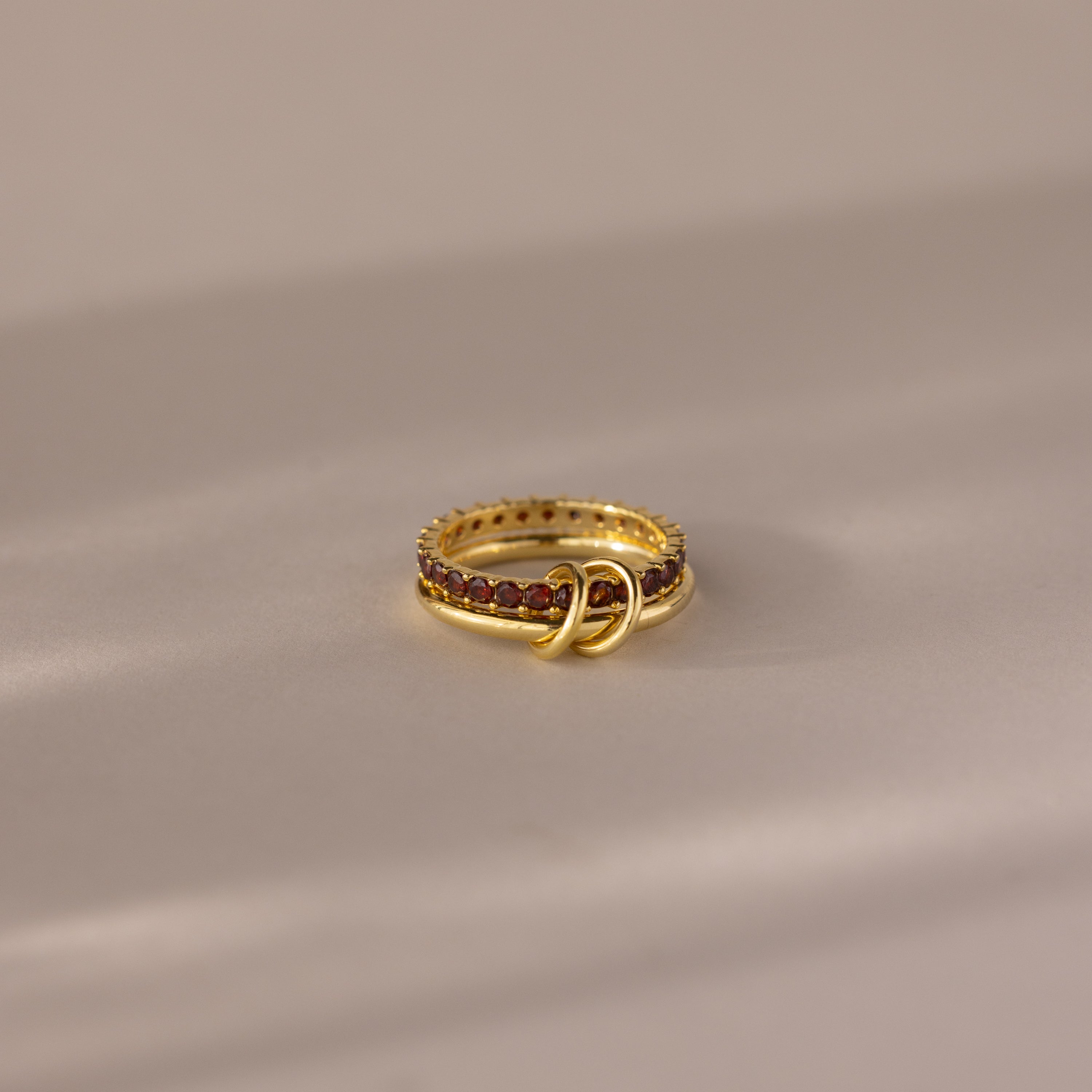 The Duo Birthstone Tennis Linked Ring Set in Garnet (January) features a minimalist, looped design with red gemstones, displayed on a soft neutral surface.