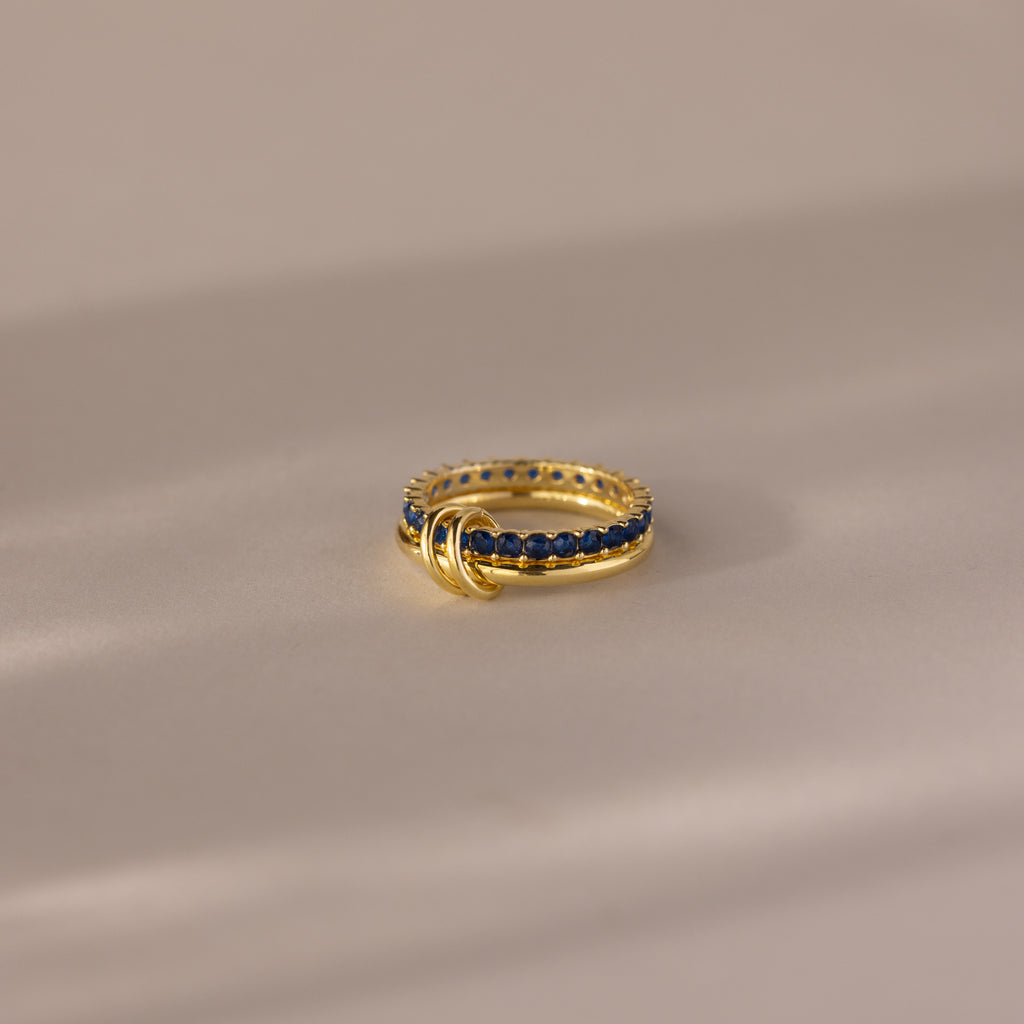 The Duo Birthstone Tennis Linked Ring Set in Sapphire (September) features a minimalist design with blue stones and three small gold loops, shown on a beige surface.