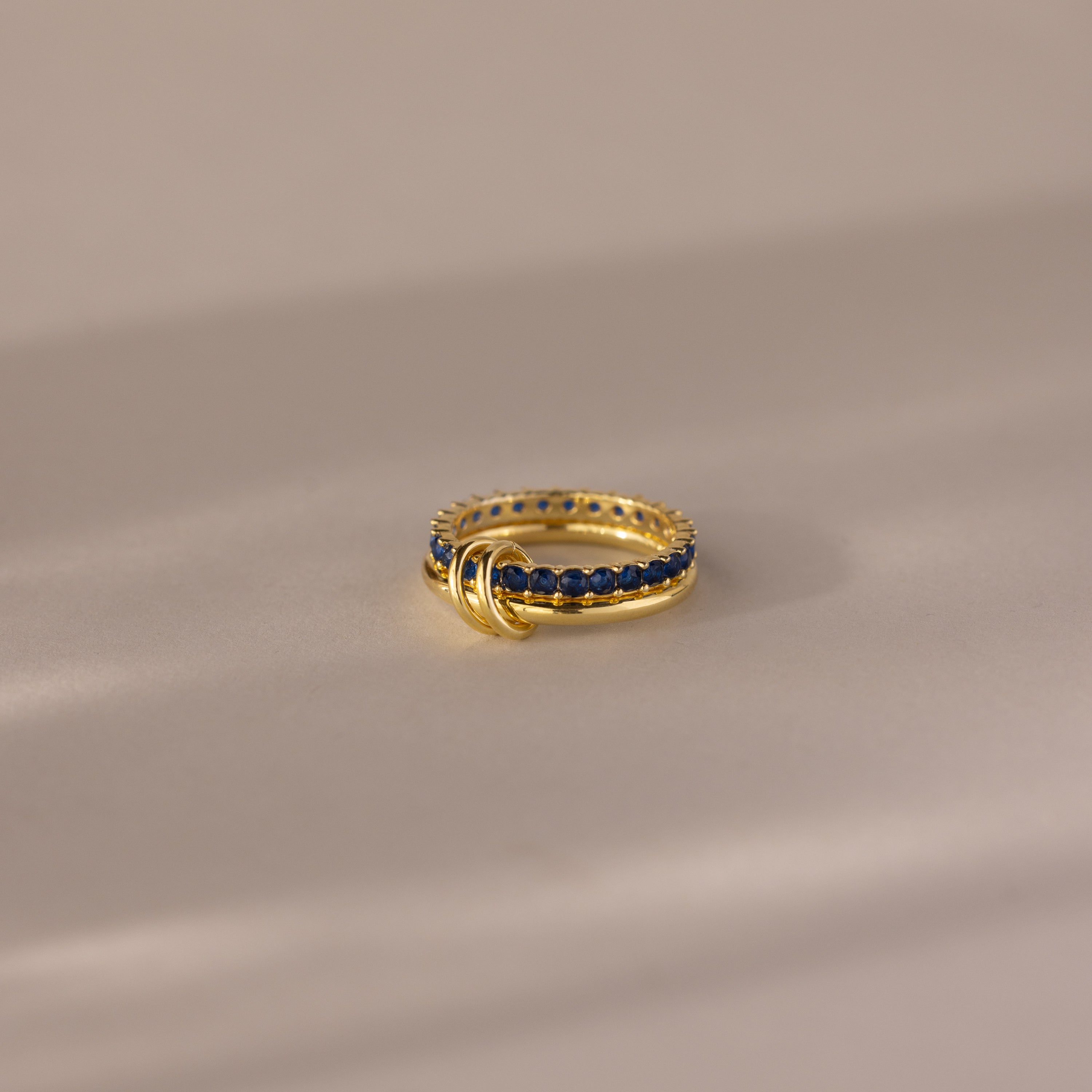 The Duo Birthstone Tennis Linked Ring Set in Sapphire (September) features a minimalist design with blue stones and three small gold loops, shown on a beige surface.