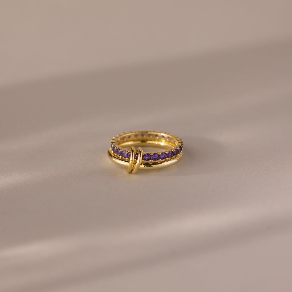 The Duo Birthstone Tennis Linked Ring Set in Amethyst (February) features minimalist gold loops and small purple stones, displayed on a smooth beige surface.