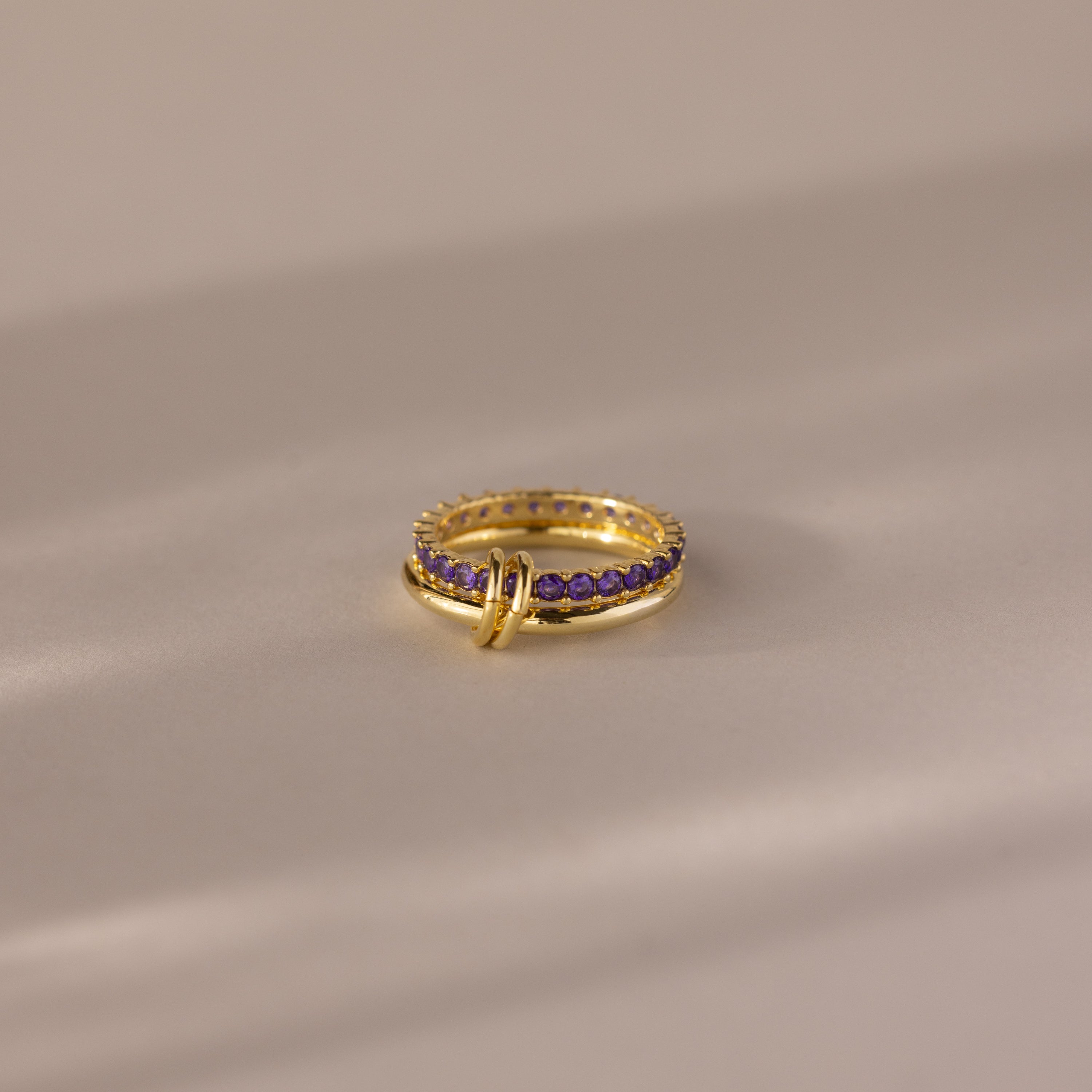 The Duo Birthstone Tennis Linked Ring Set in Amethyst (February) features minimalist gold loops and small purple stones, displayed on a smooth beige surface.