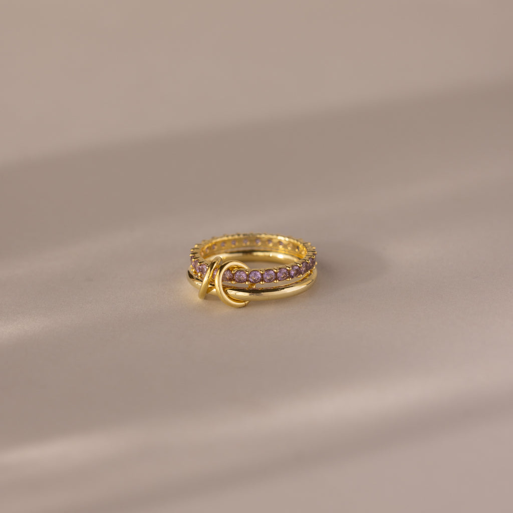 The Duo Alexandrite Tennis Linked Ring Set features small pink stones and a double loop design, elegantly displayed on a softly lit, neutral surface.