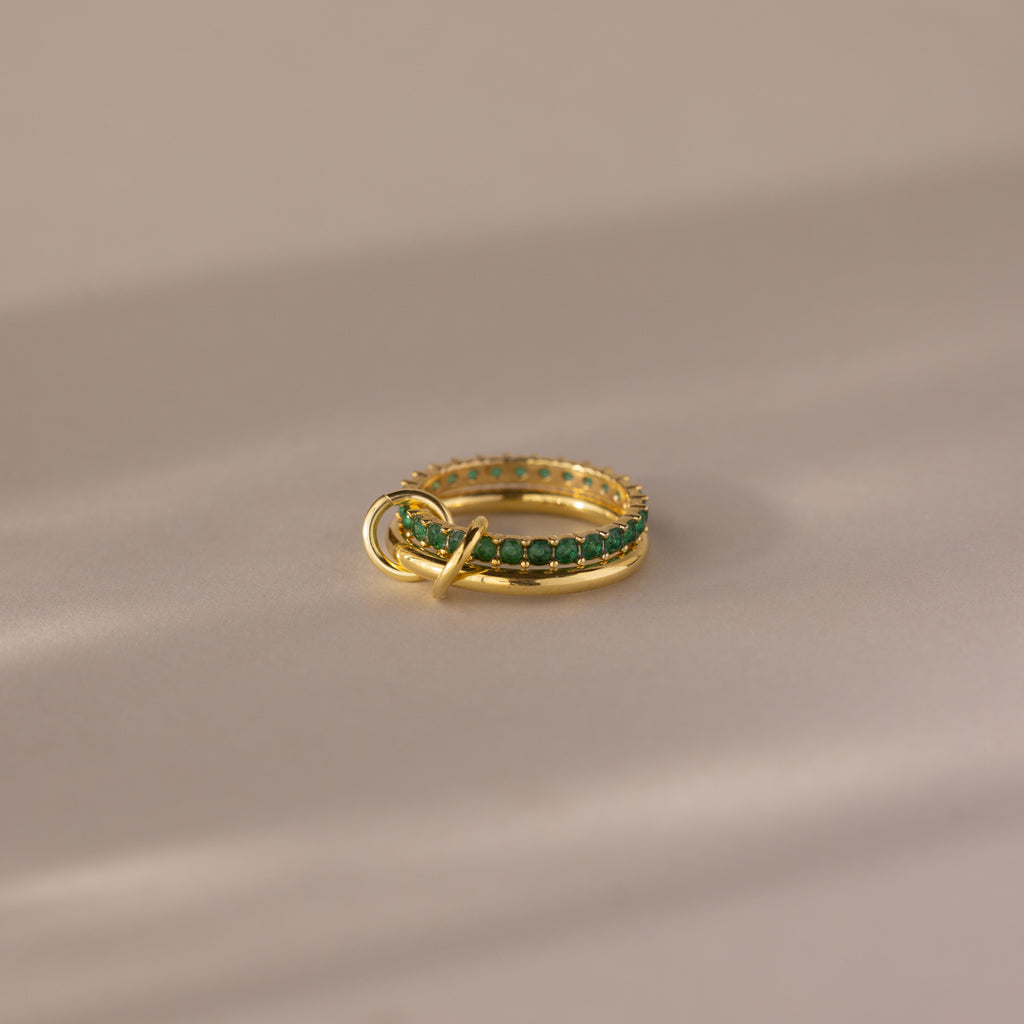 The Duo Birthstone Tennis Linked Ring Set in emerald (May) is displayed on a beige, softly lit surface.