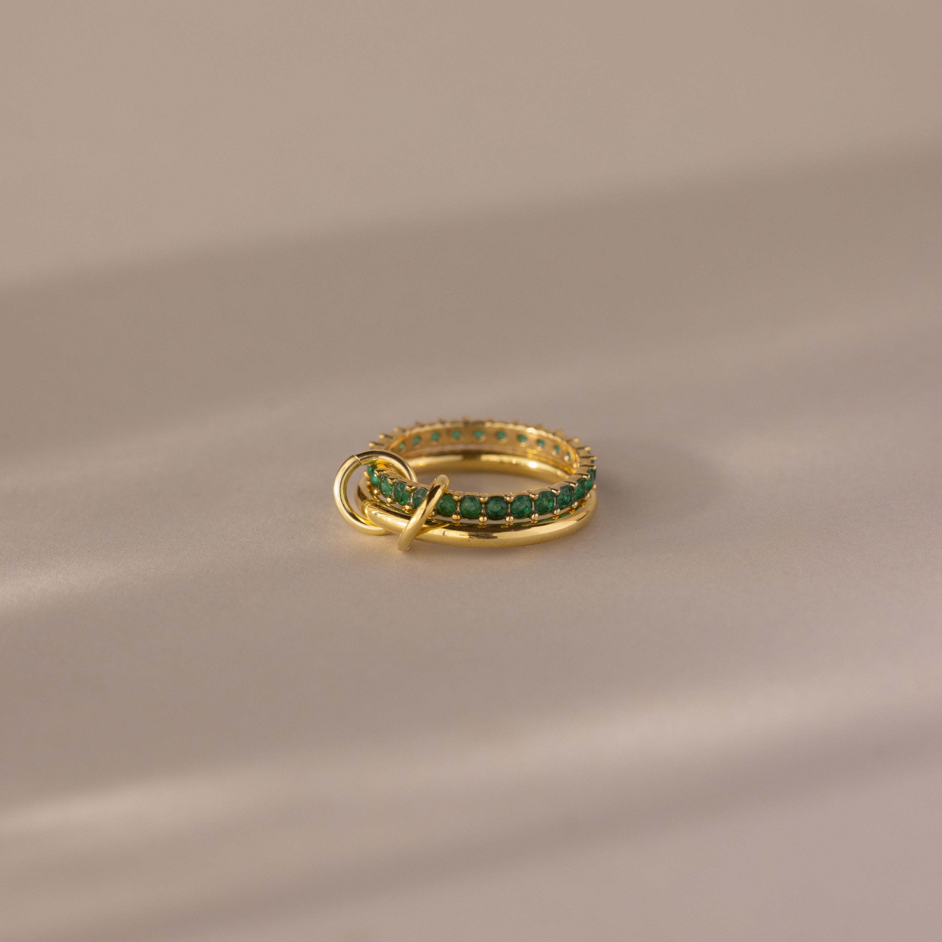 The Duo Birthstone Tennis Linked Ring Set in emerald (May) is displayed on a beige, softly lit surface.