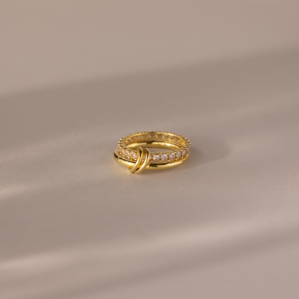 Duo Birthstone Tennis Linked Ring Set in Diamond (April) features two interlocking gold loops with small diamonds, displayed on a smooth, neutral surface.
