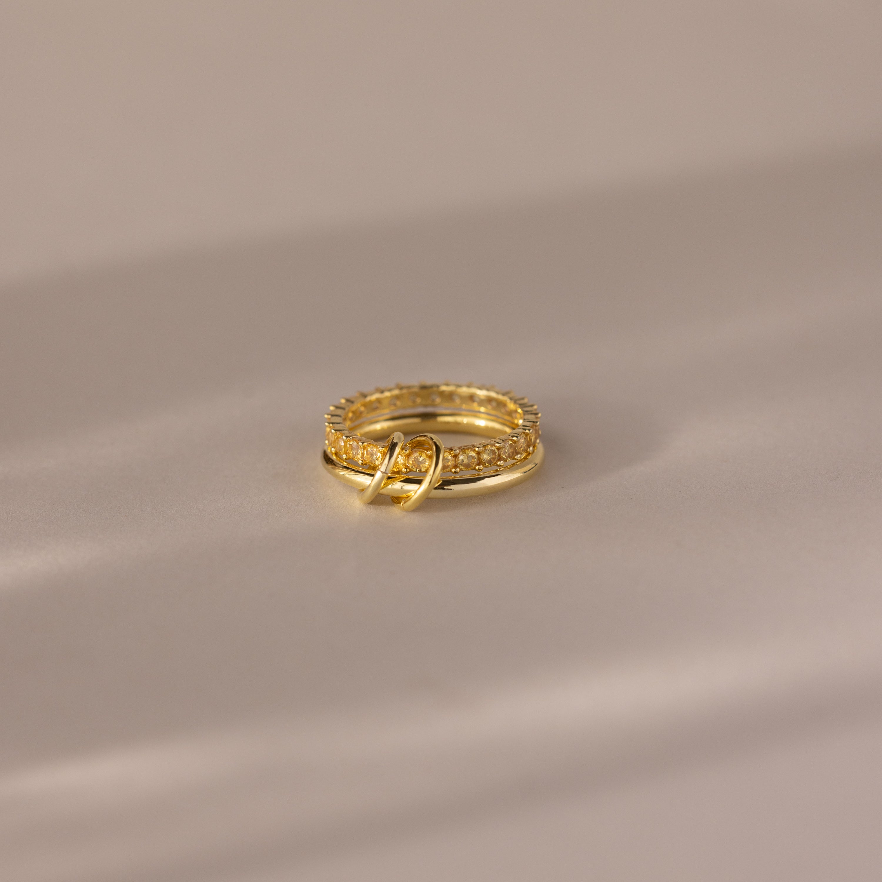 The Duo Birthstone Tennis Linked Ring Set in Topaz (November)—a minimalist gold ring with small clear stones intertwined with a plain gold band—rests on a beige surface.