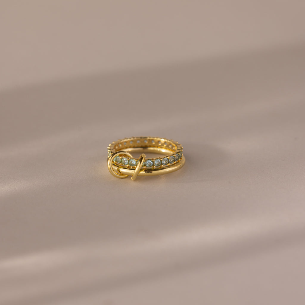 The Duo Birthstone Tennis Linked Ring Set in Aquamarine (March) features a minimalist gold band with blue stones, artfully displayed on a soft beige surface.