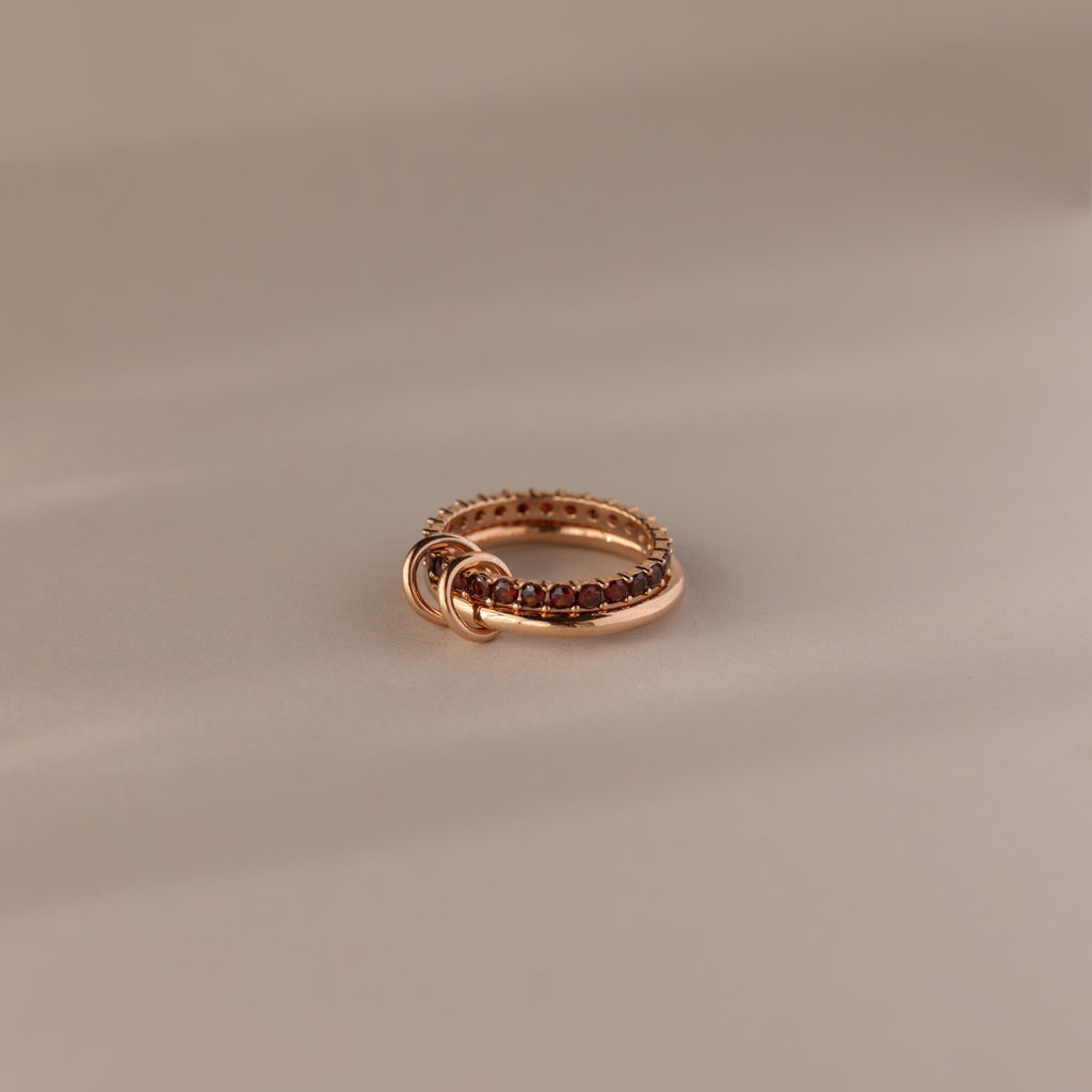 The Duo Garnet Tennis Linked Ring Set showcases small red gemstones set around the band, elegantly displayed on a beige surface.