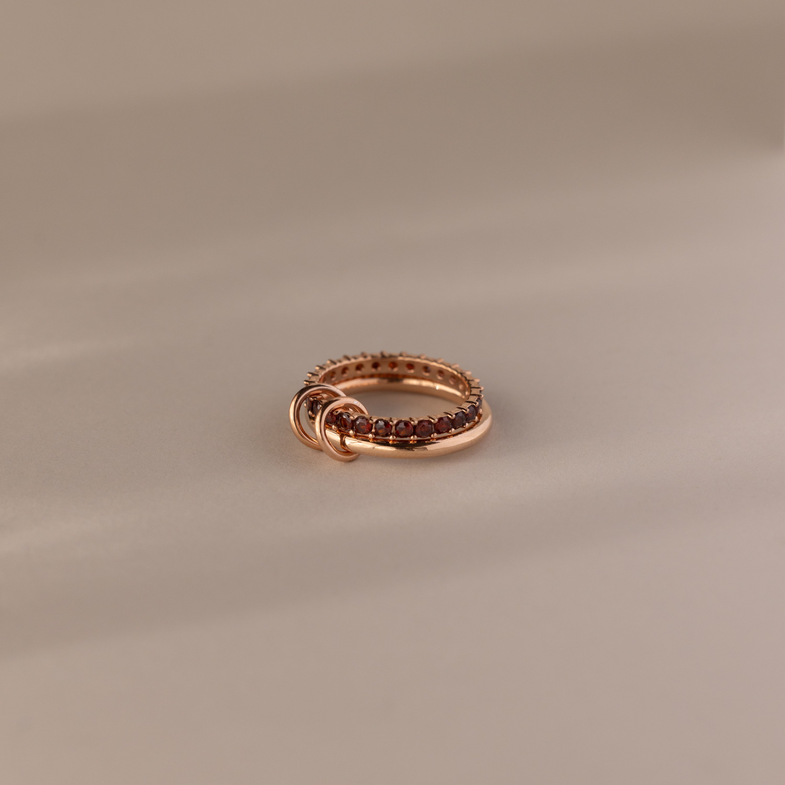 The Duo Garnet Tennis Linked Ring Set showcases small red gemstones set around the band, elegantly displayed on a beige surface.
