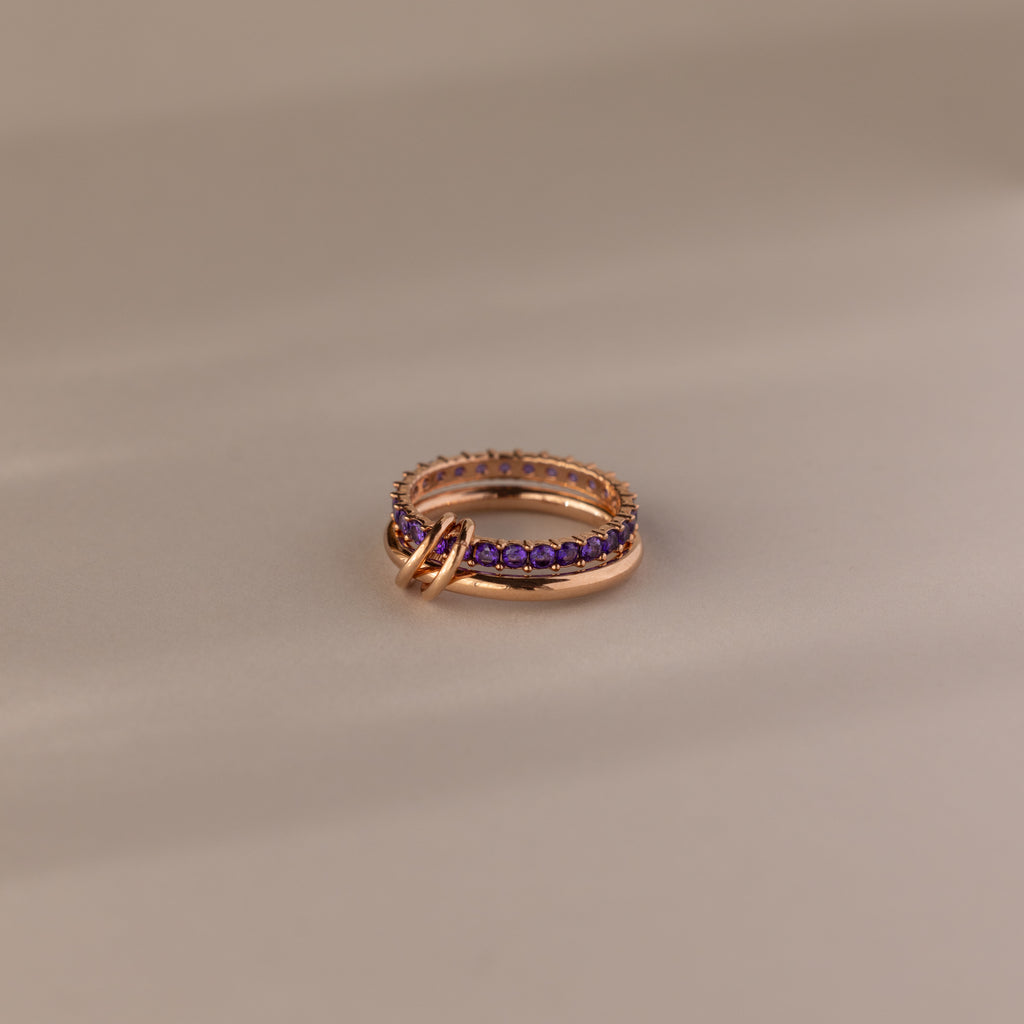 The Duo Amethyst Tennis Linked Ring Set, featuring purple amethyst gemstones and interlocking gold rings, is elegantly displayed on a beige surface.