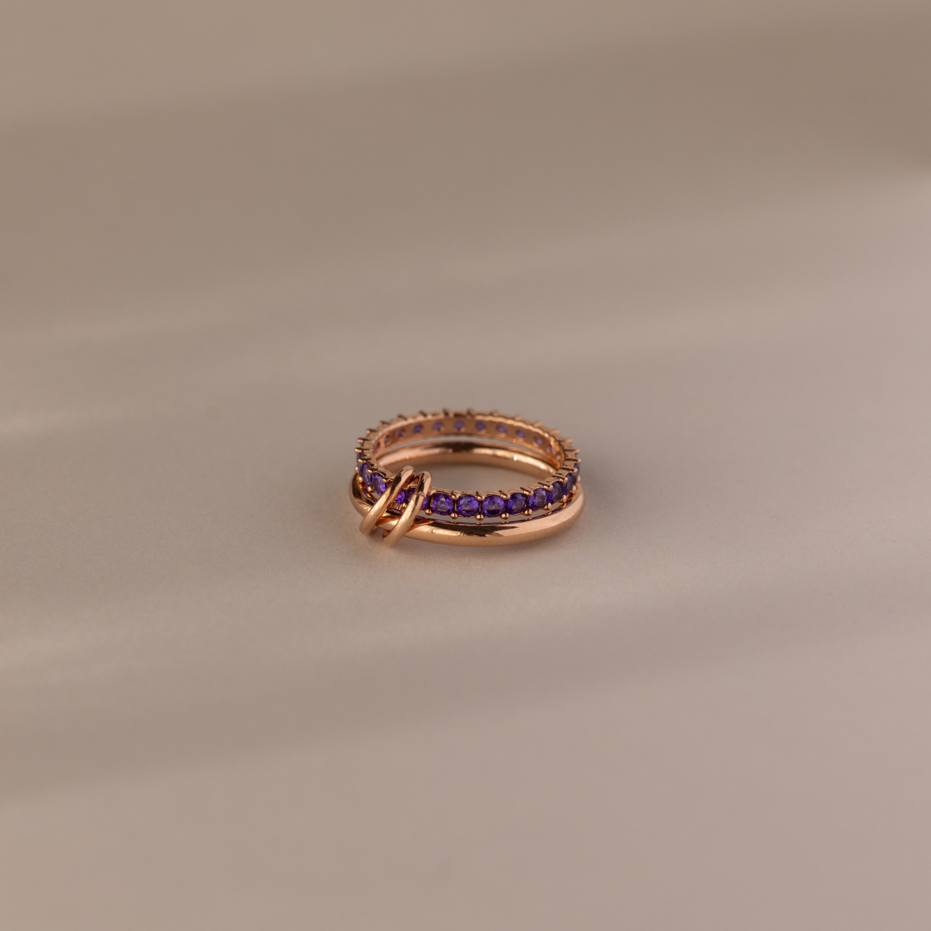 The Duo Amethyst Tennis Linked Ring Set, featuring purple amethyst gemstones and interlocking gold rings, is elegantly displayed on a beige surface.