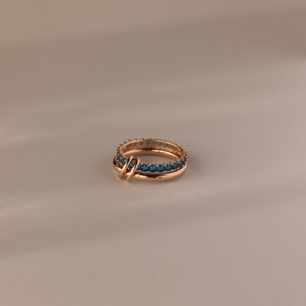 The Duo Blue Zircon Tennis Linked Ring Set features shimmering blue zircon stones in a row, showcased on a neutral beige surface.