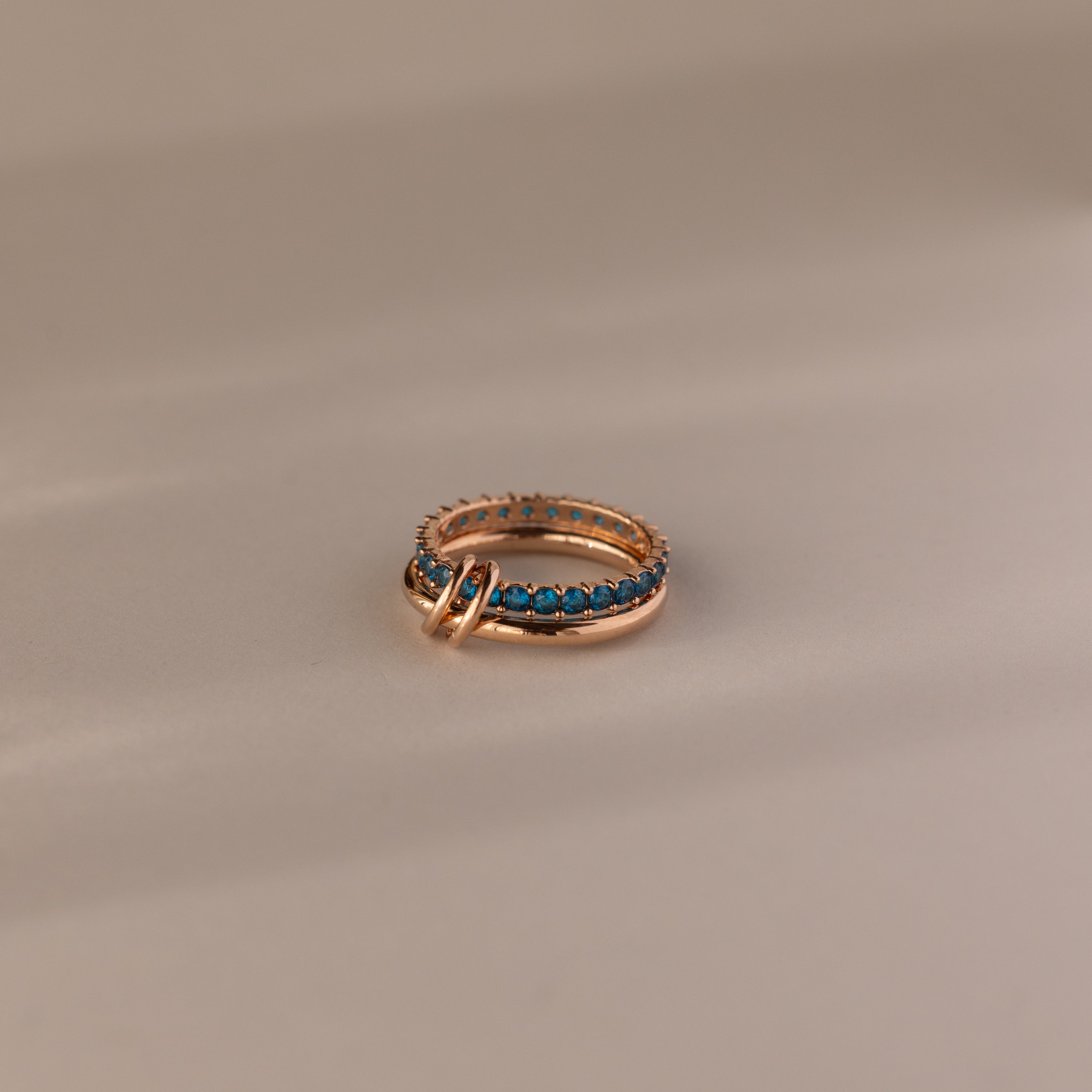 The Duo Blue Zircon Tennis Linked Ring Set features shimmering blue zircon stones in a row, showcased on a neutral beige surface.