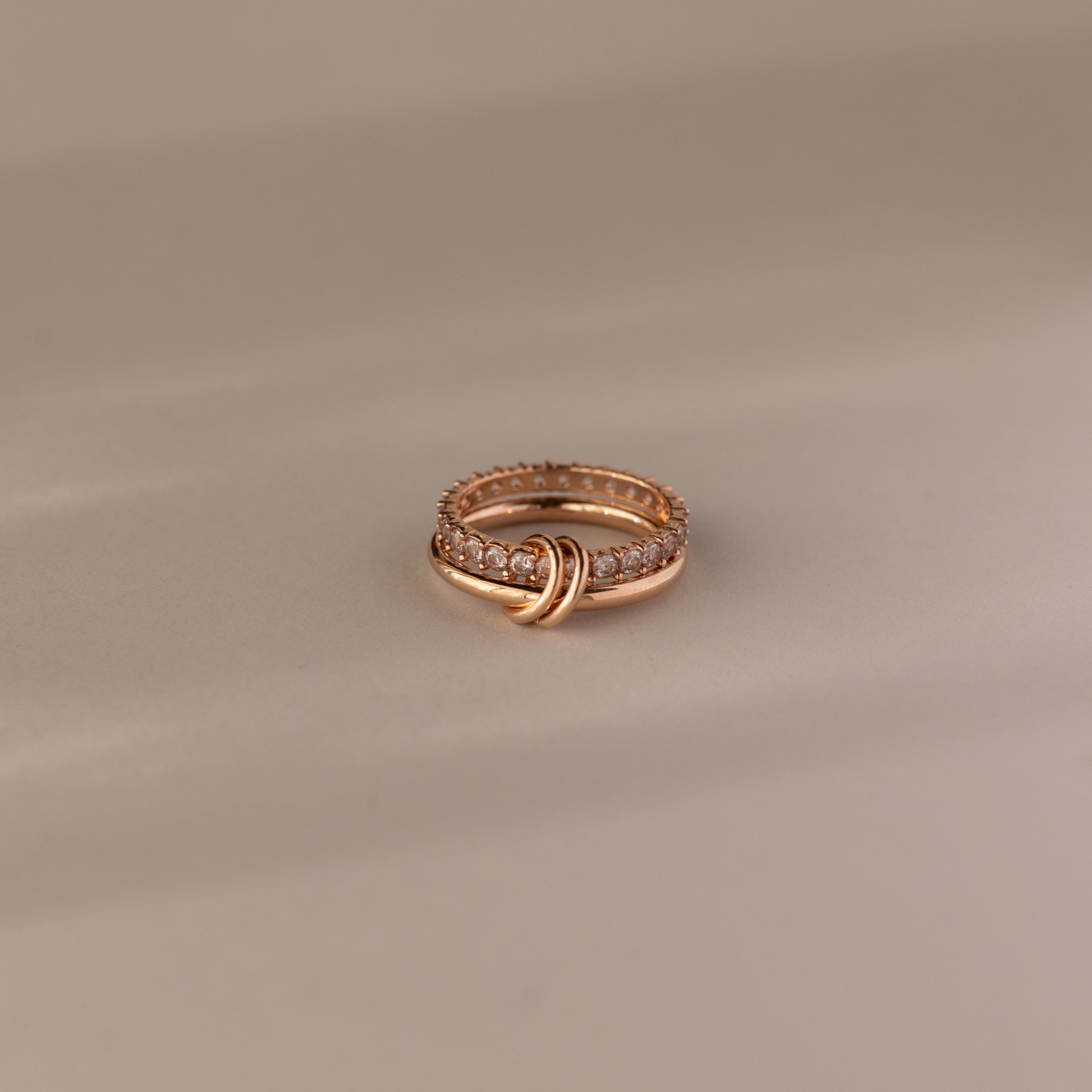 The Duo Diamond Tennis Linked Ring Set showcases a minimalist gold knot design with sparkling diamonds, elegantly displayed on a soft beige surface.