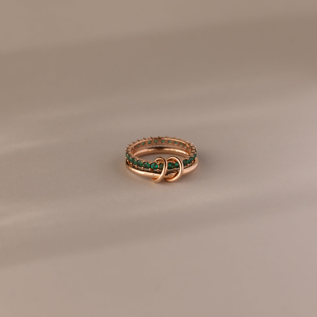 The Duo Emerald Tennis Linked Ring Set features two interlocking gold bands adorned with green gemstones, creating a unique minimalist birthstone ring against a neutral beige background.