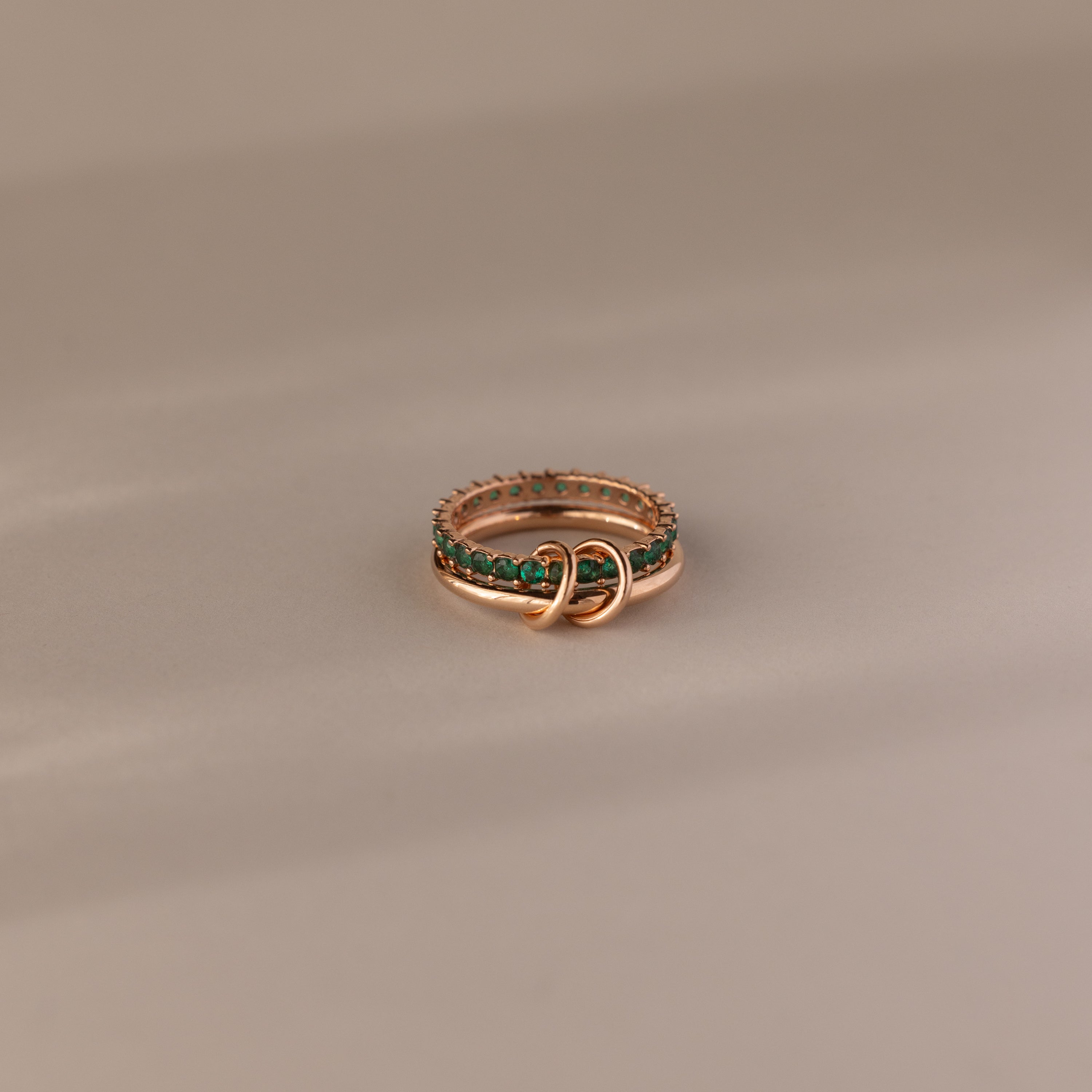 The Duo Emerald Tennis Linked Ring Set features two interlocking gold bands adorned with green gemstones, creating a unique minimalist birthstone ring against a neutral beige background.