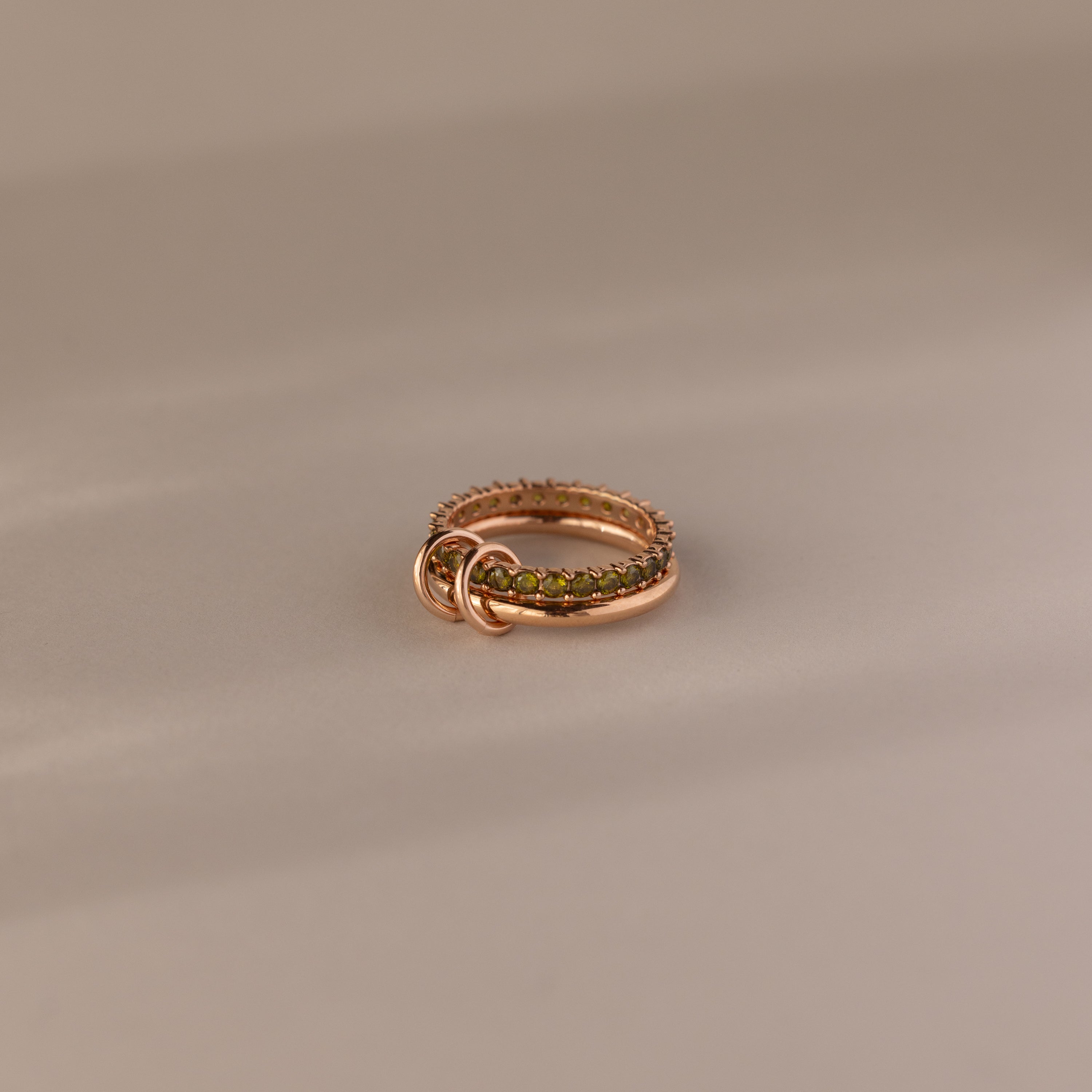 The Duo Peridot Tennis Linked Ring Set features green gemstones and two gold loops, displayed on a beige surface.