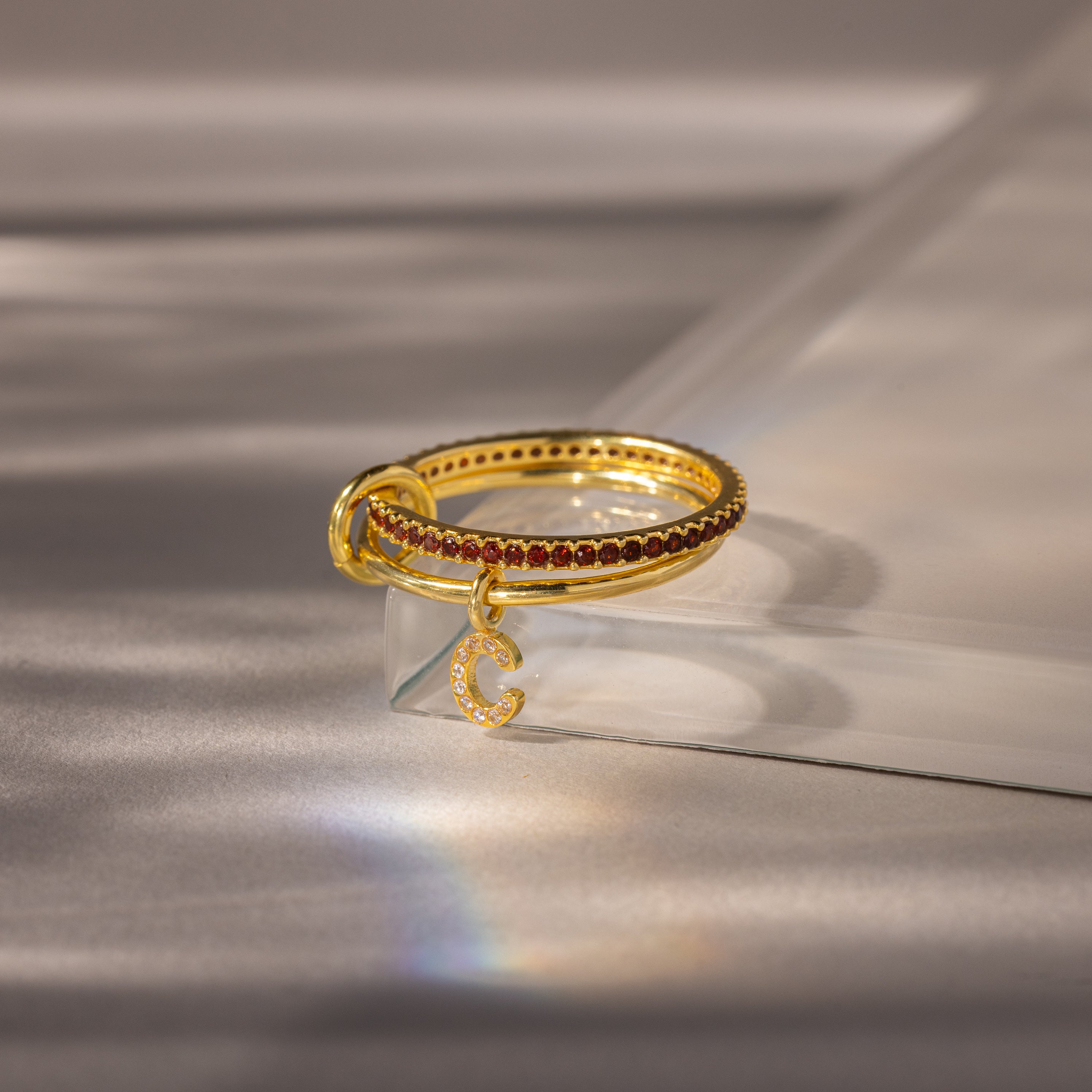 Linked Initial Garnet Ring Set features small red gemstones and a crescent moon charm, displayed on a clear acrylic stand—personalized jewelry perfect as a birthstone ring or unique accessory.