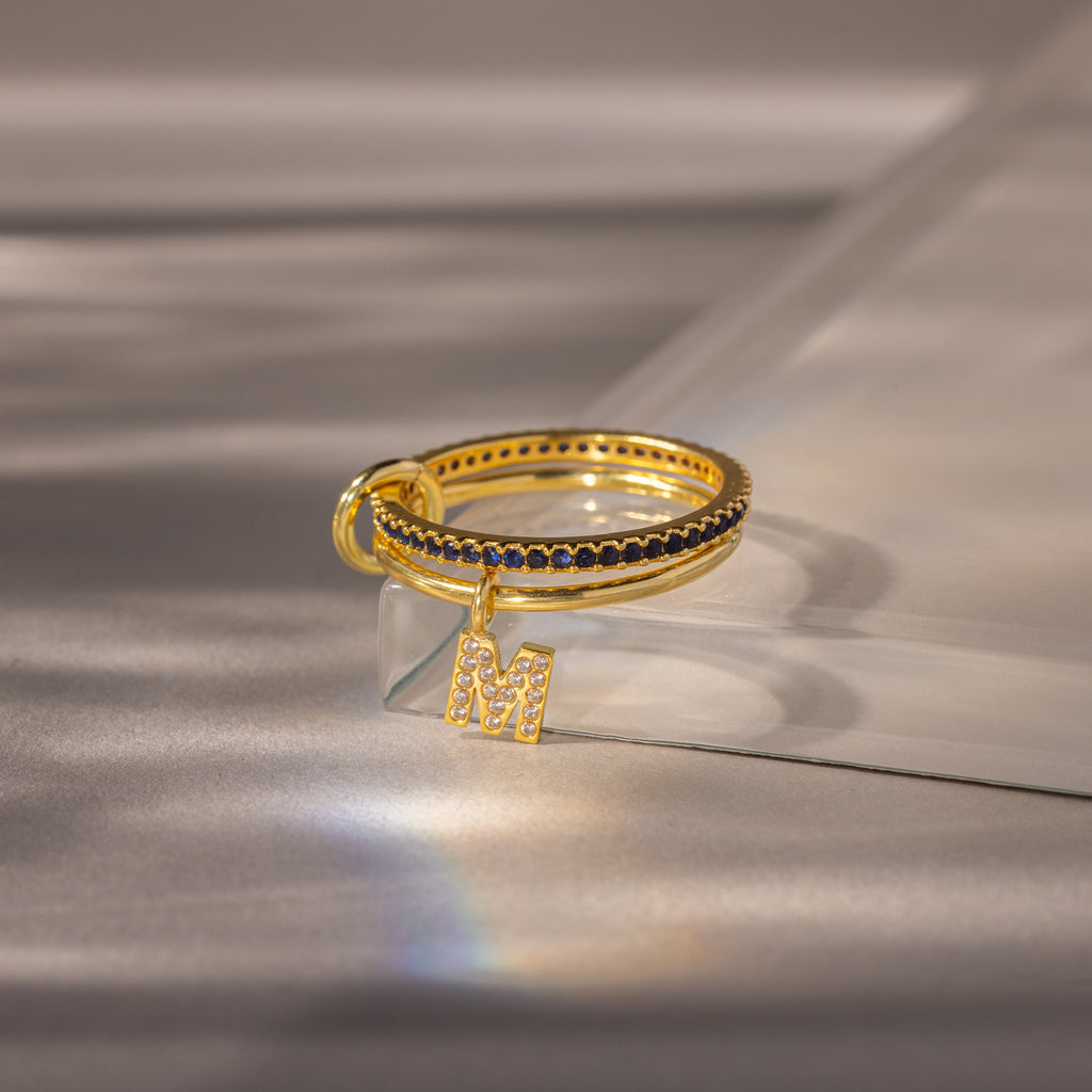 The Linked Initial Sapphire Ring Set features a gold ring with black stones and a sparkling letter M charm, adding personalized elegance to any style, beautifully displayed on a soft reflective surface.