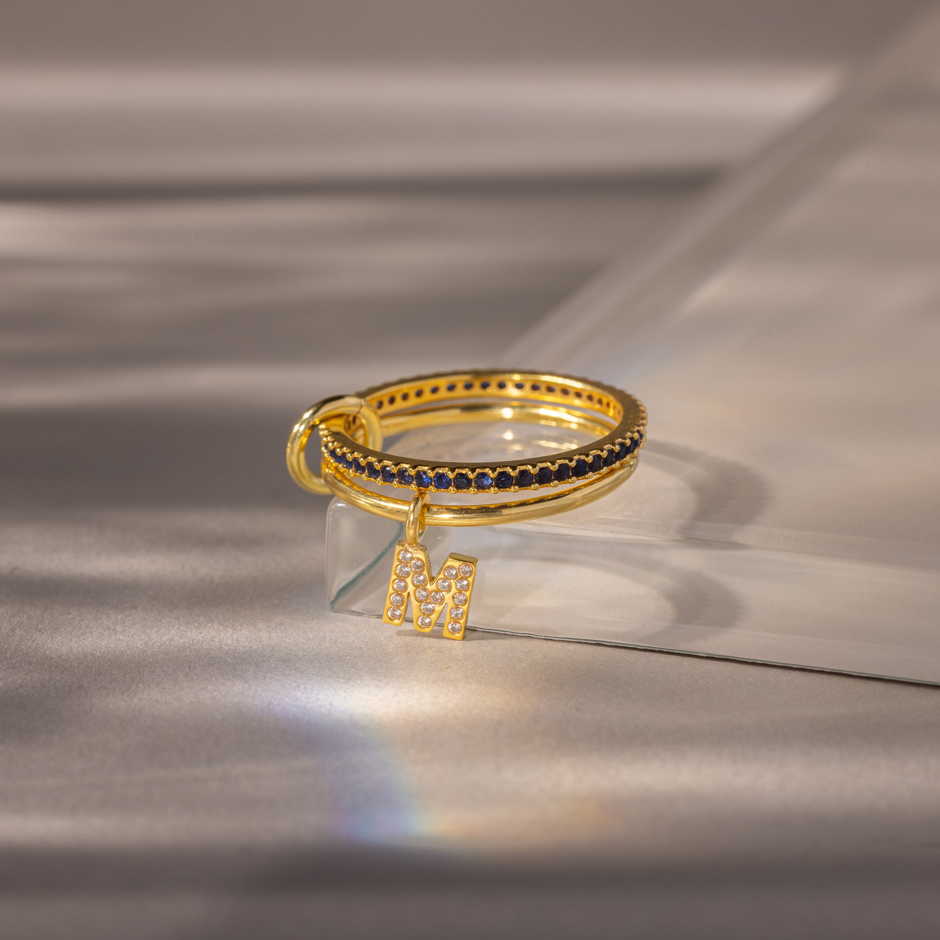 The Linked Initial Sapphire Ring Set features a gold ring with black stones and a sparkling letter M charm, adding personalized elegance to any style, beautifully displayed on a soft reflective surface.