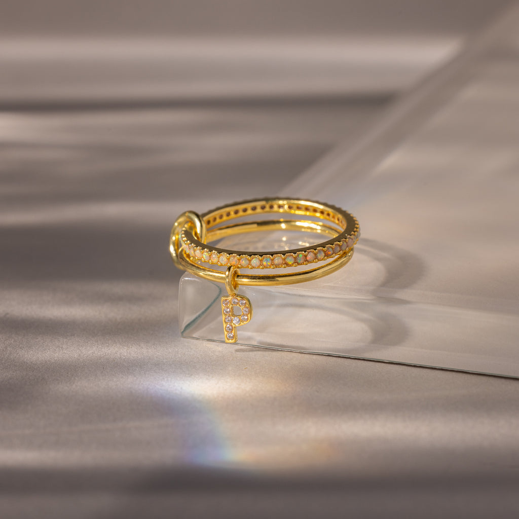 Linked Initial Opal Ring Set featuring sparkling accents and a dangling letter P charm, displayed on a clear acrylic surface.