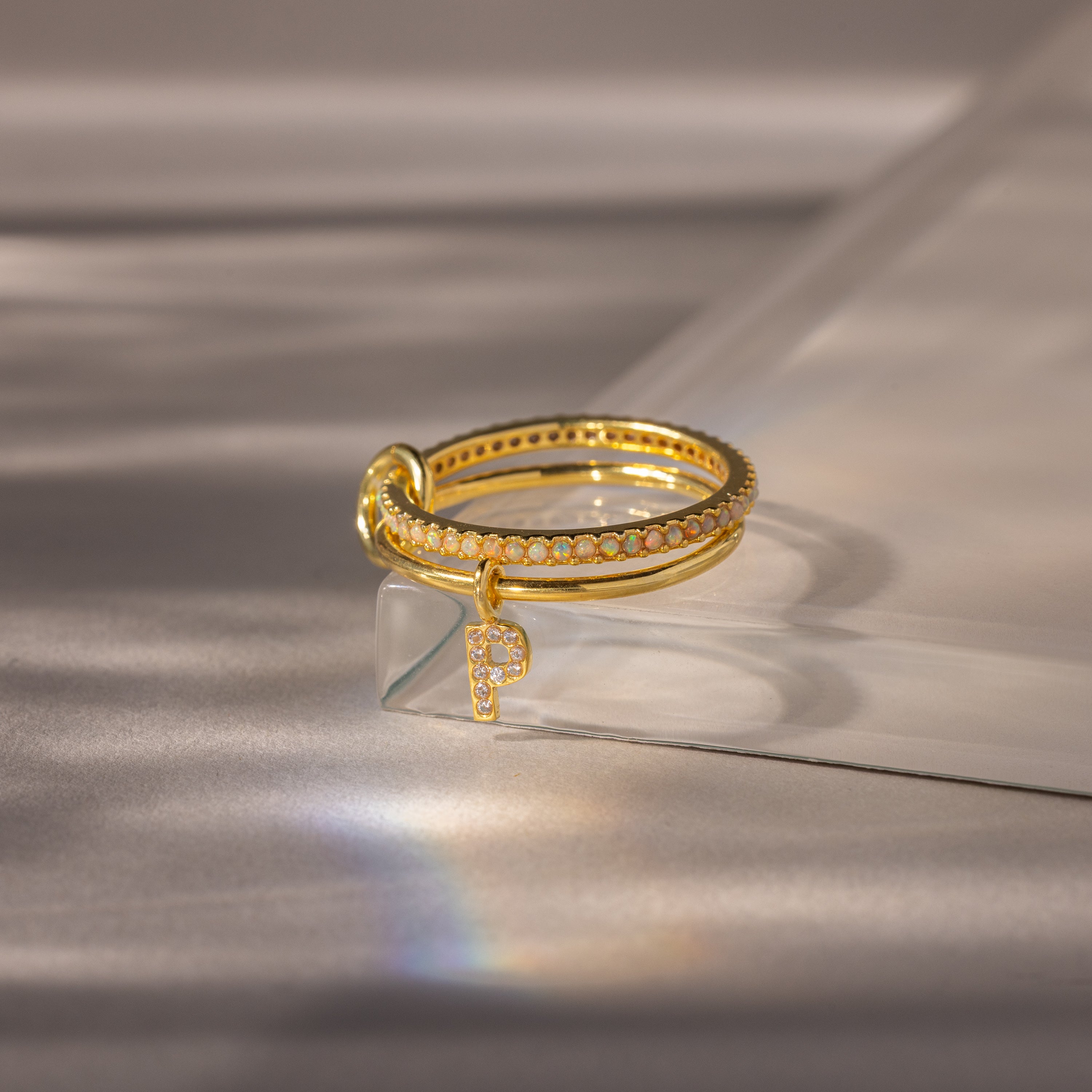 Linked Initial Opal Ring Set featuring sparkling accents and a dangling letter P charm, displayed on a clear acrylic surface.