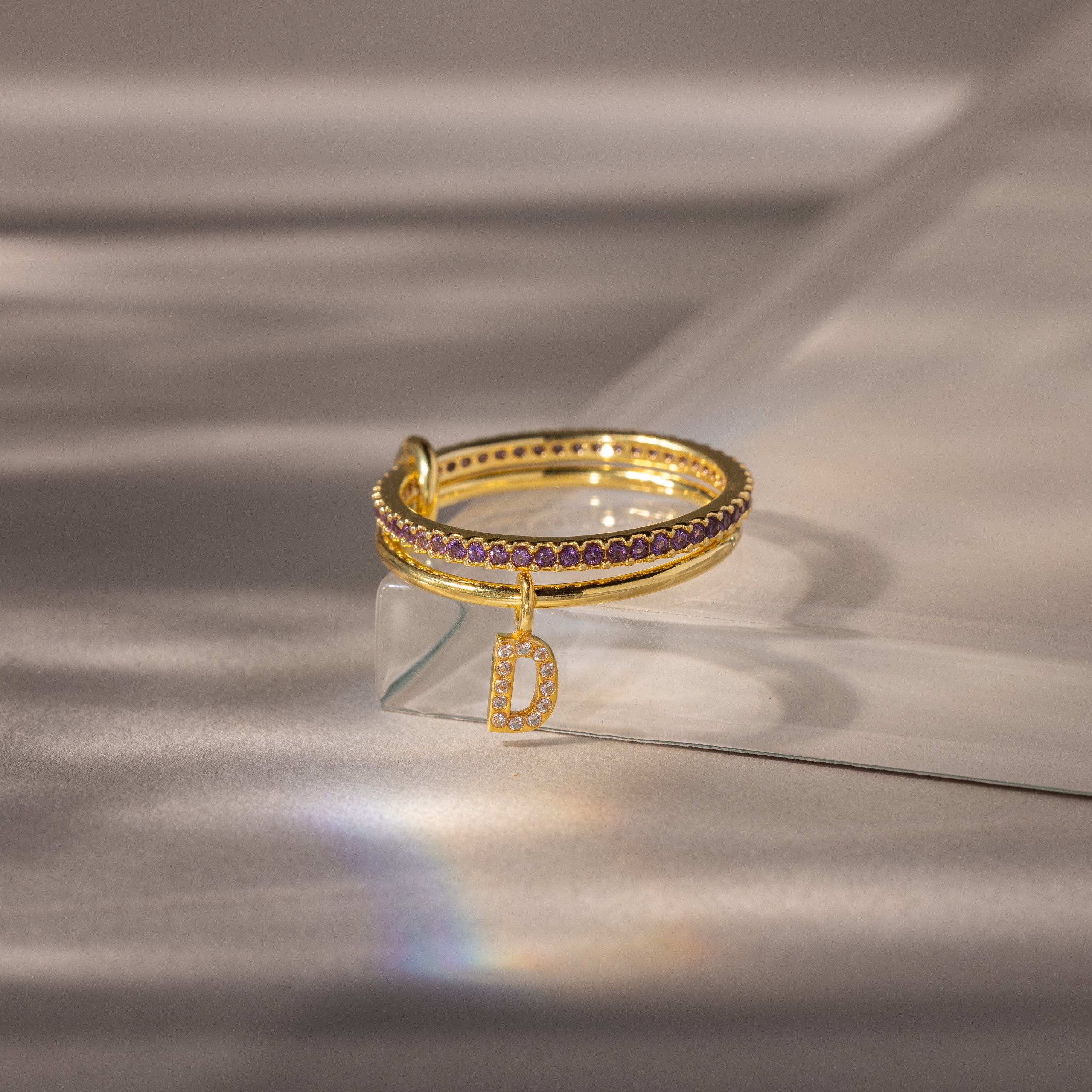 Linked Initial Birthstone Ring Set in Amethyst (February), featuring purple stones and a dangling letter D charm, displayed on a reflective surface.