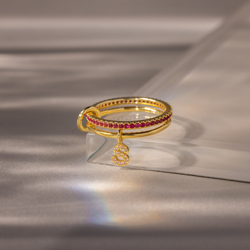 Linked Initial Ruby Ring Set with red gemstones and a dangling pavé initial charm, showcased on a light surface with soft shadows.