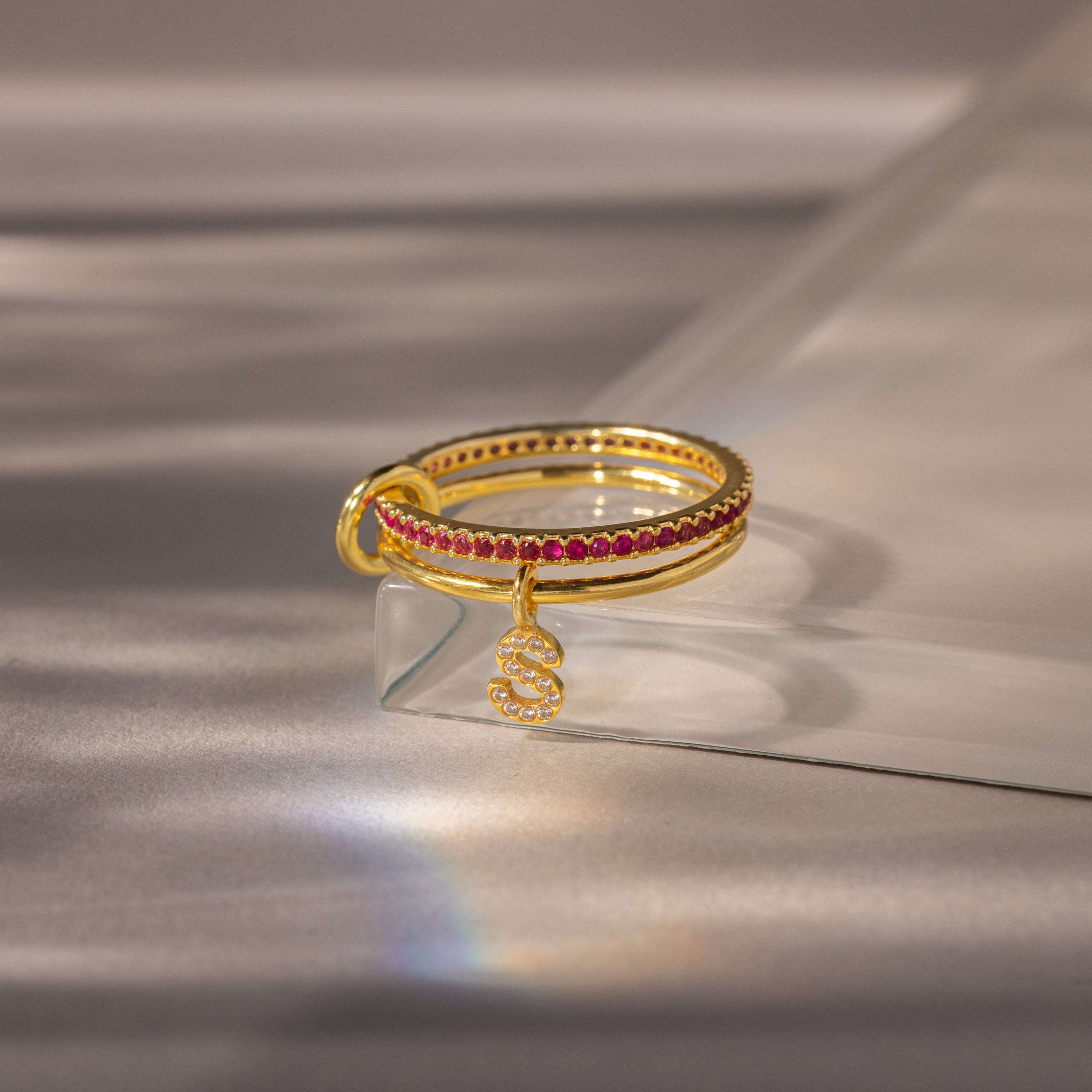 Linked Initial Ruby Ring Set with red gemstones and a dangling pavé initial charm, showcased on a light surface with soft shadows.