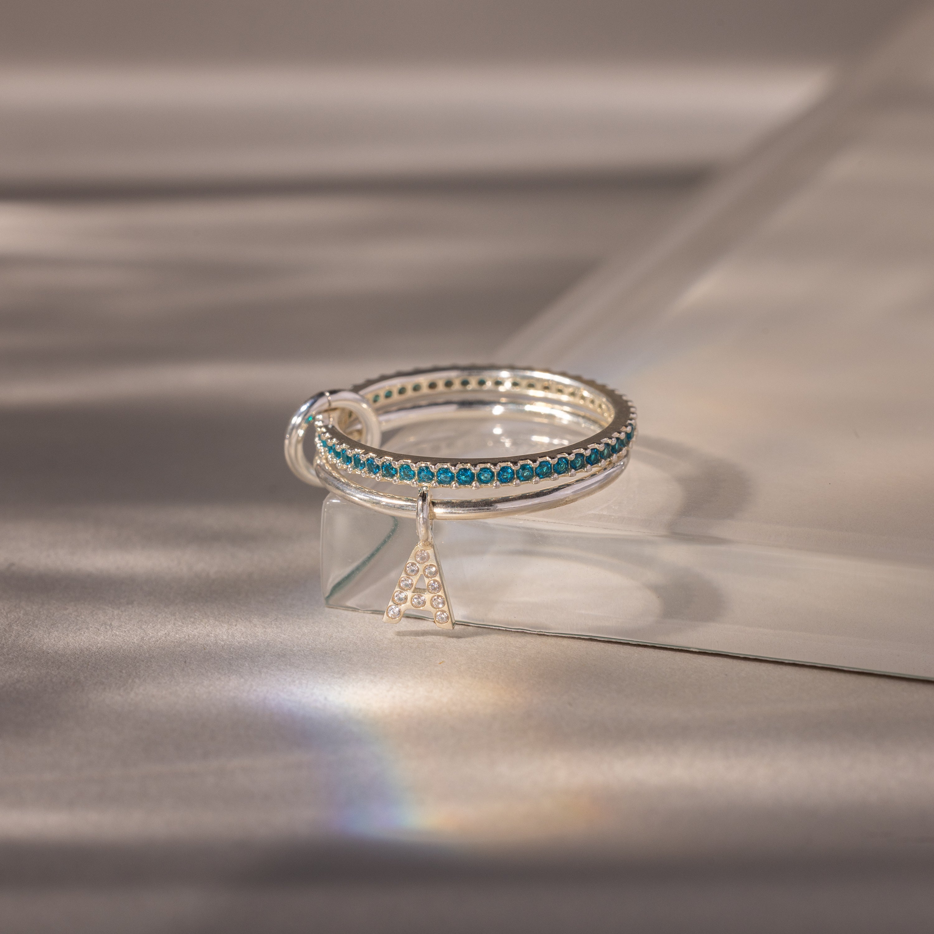 The Linked Initial Blue Zircon Ring Set showcases blue stones and a diamond-studded A charm, elegantly displayed on a glass surface—a perfect choice for personalized jewelry.