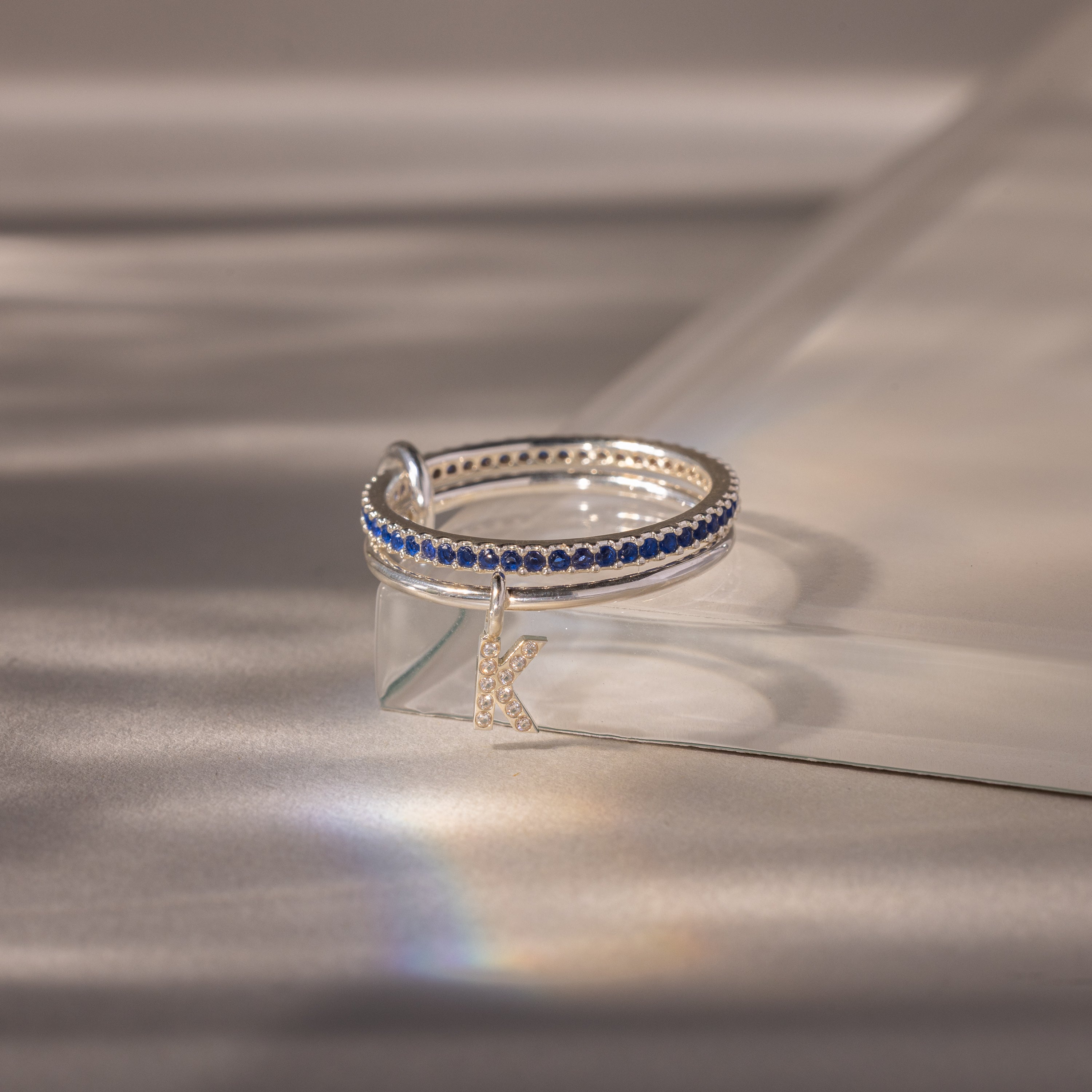 The Linked Initial Sapphire Ring Set, with blue stones and a K charm, is displayed on a clear angled surface in soft light—ideal as personalized jewelry or a distinctive birthstone ring.