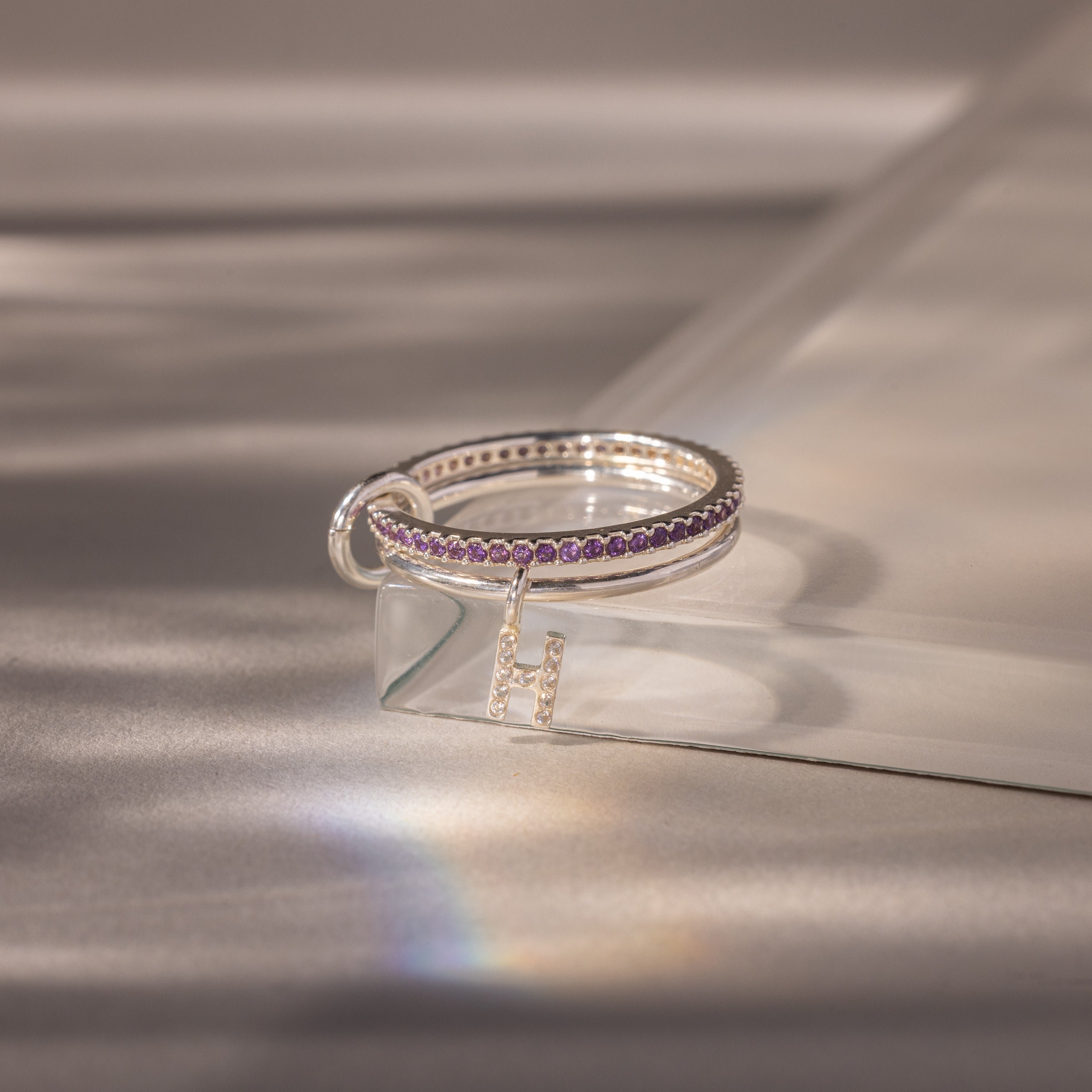 The Linked Initial Amethyst Ring Set features silver rings with purple amethyst stones and an "H" initial charm, displayed on a clear, angled surface.