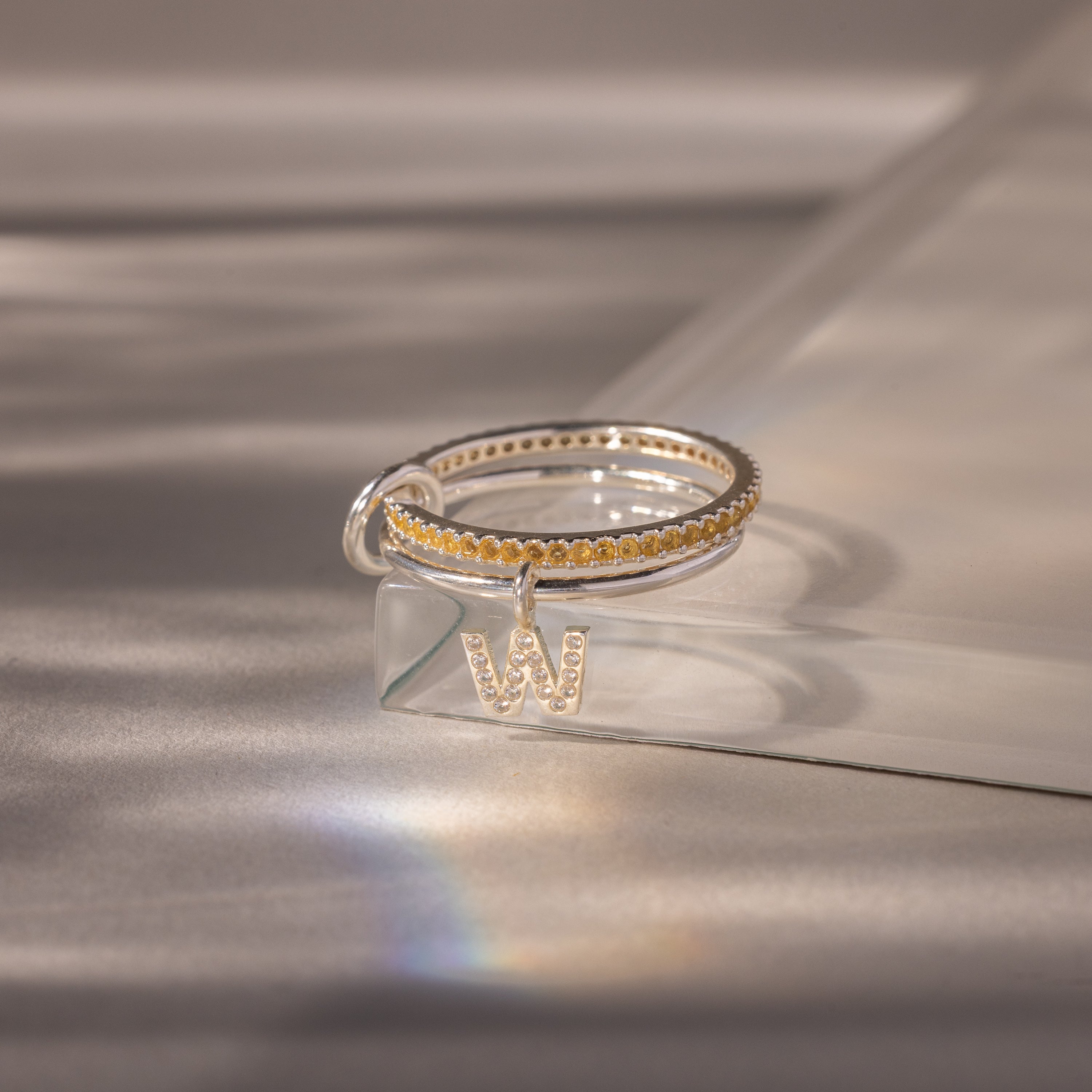 The Linked Initial Topaz Ring Set features yellow stones and a pavé initial charm, beautifully presented on a soft, reflective surface.