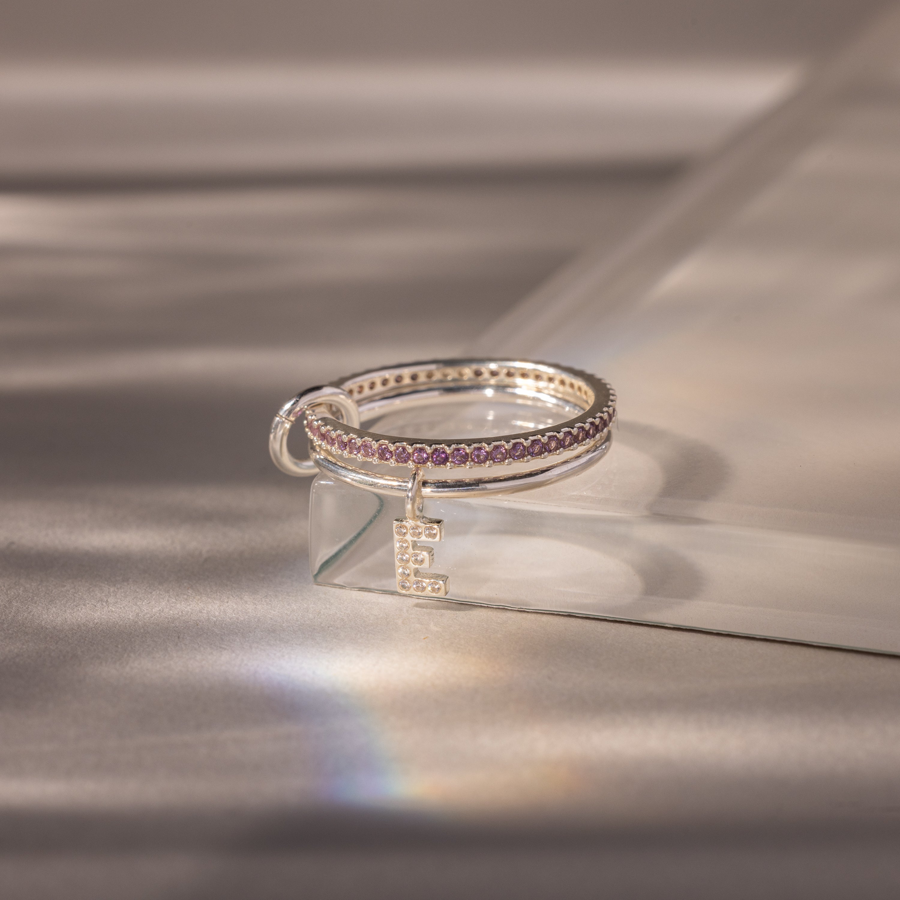 The Linked Initial Alexandrite Ring Set features a dazzling E charm and purple stones, elegantly showcased on clear glass under soft lighting.