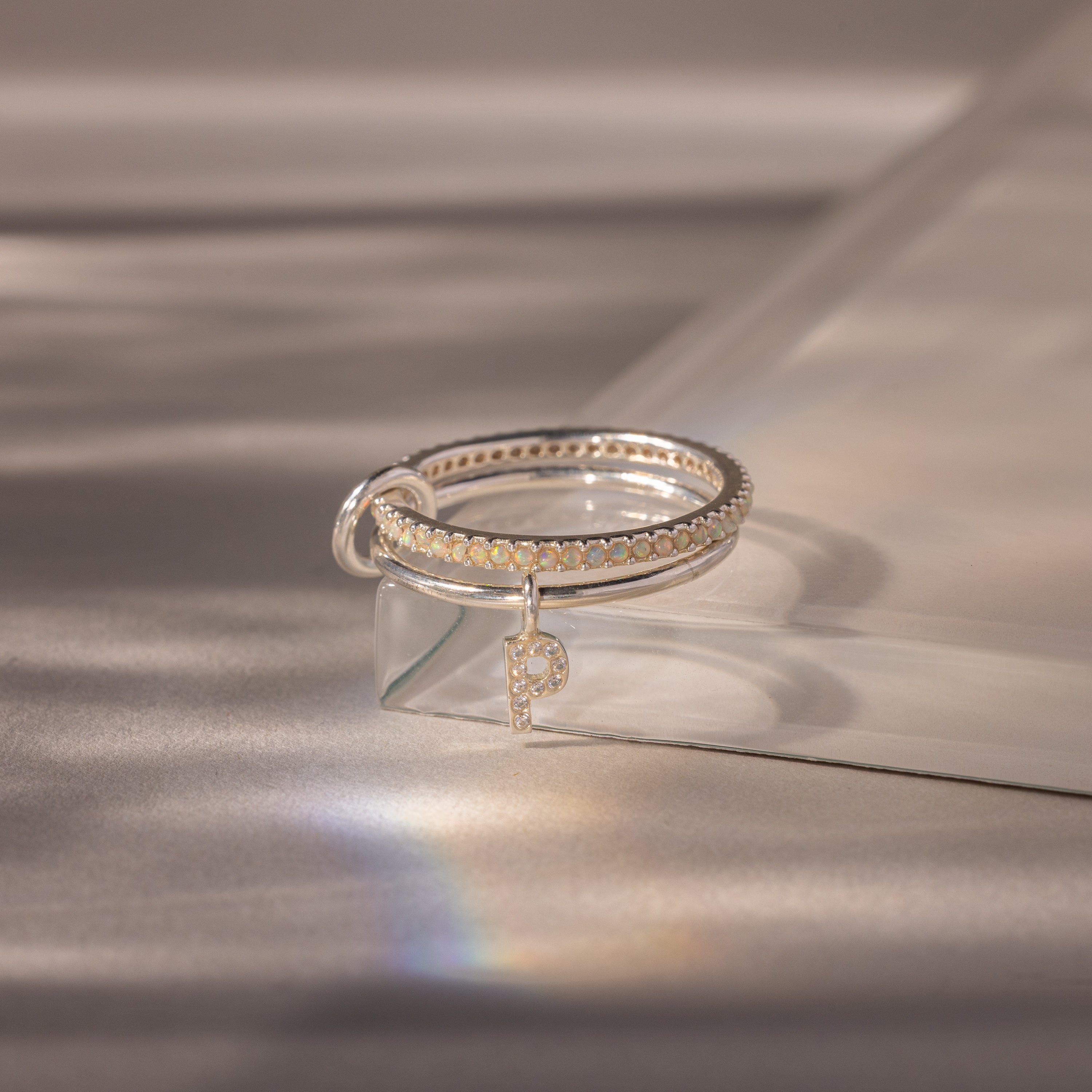 The Linked Initial Opal Ring Set, featuring a silver ring with an initial pendant, is displayed on a glass surface under gentle lighting.