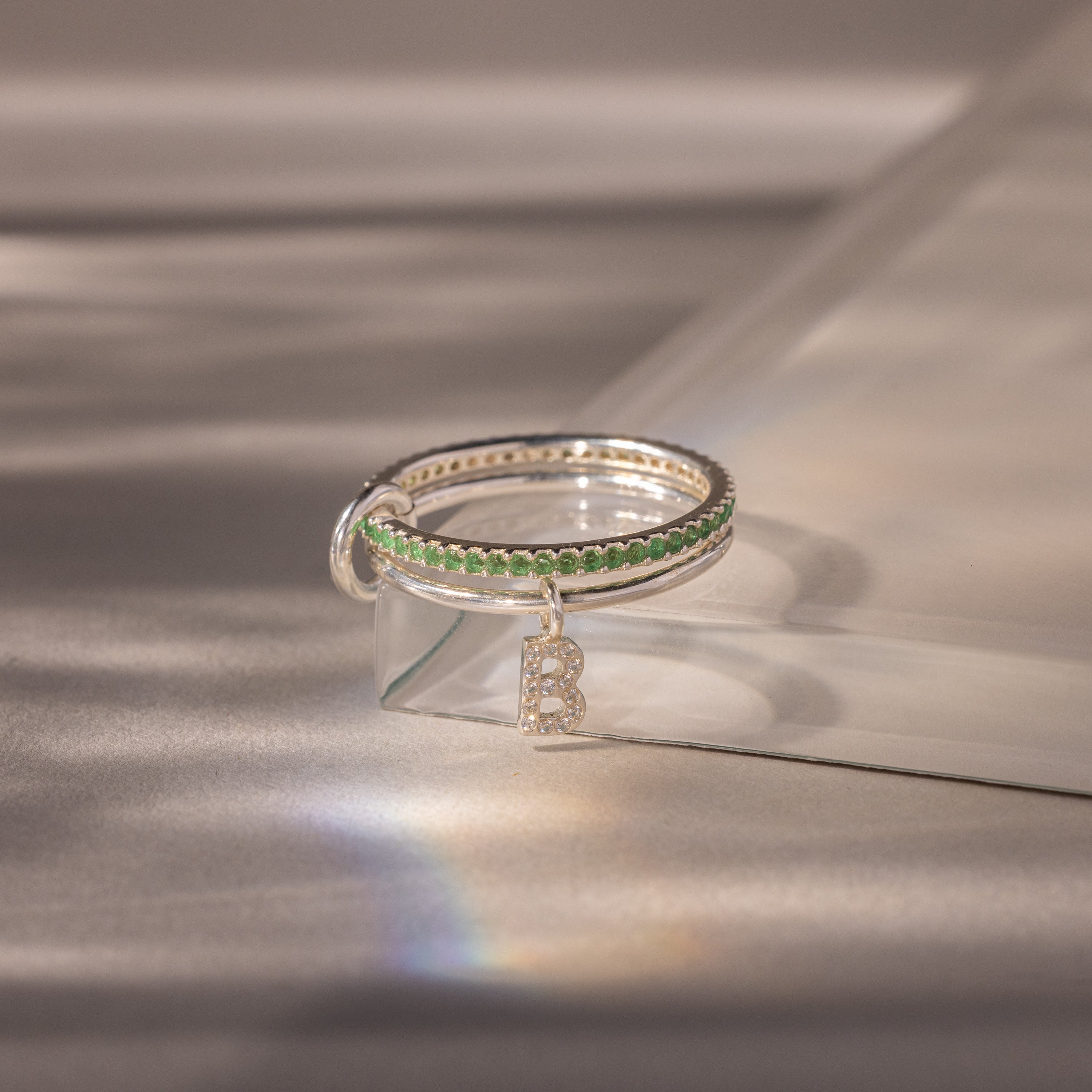 The Linked Initial Emerald Ring Set features green stones reminiscent of an Emerald Ring, a dangling letter B charm, and comes on a clear stand—ideal as Birthstone Jewelry or a unique personalized gift.