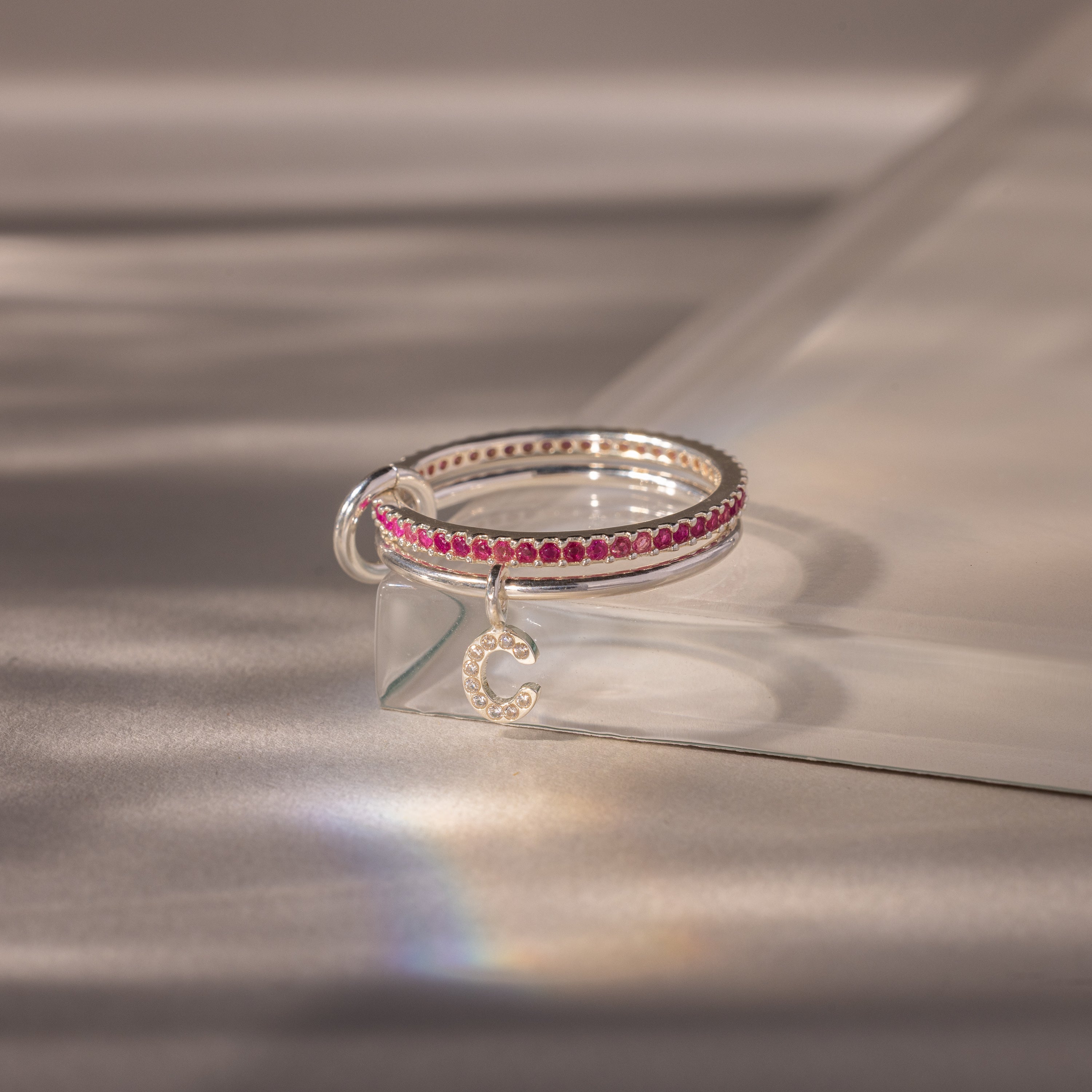 The Linked Initial Ruby Ring Set features pink gemstones and a pavé letter C charm, displayed on a clear geometric block.