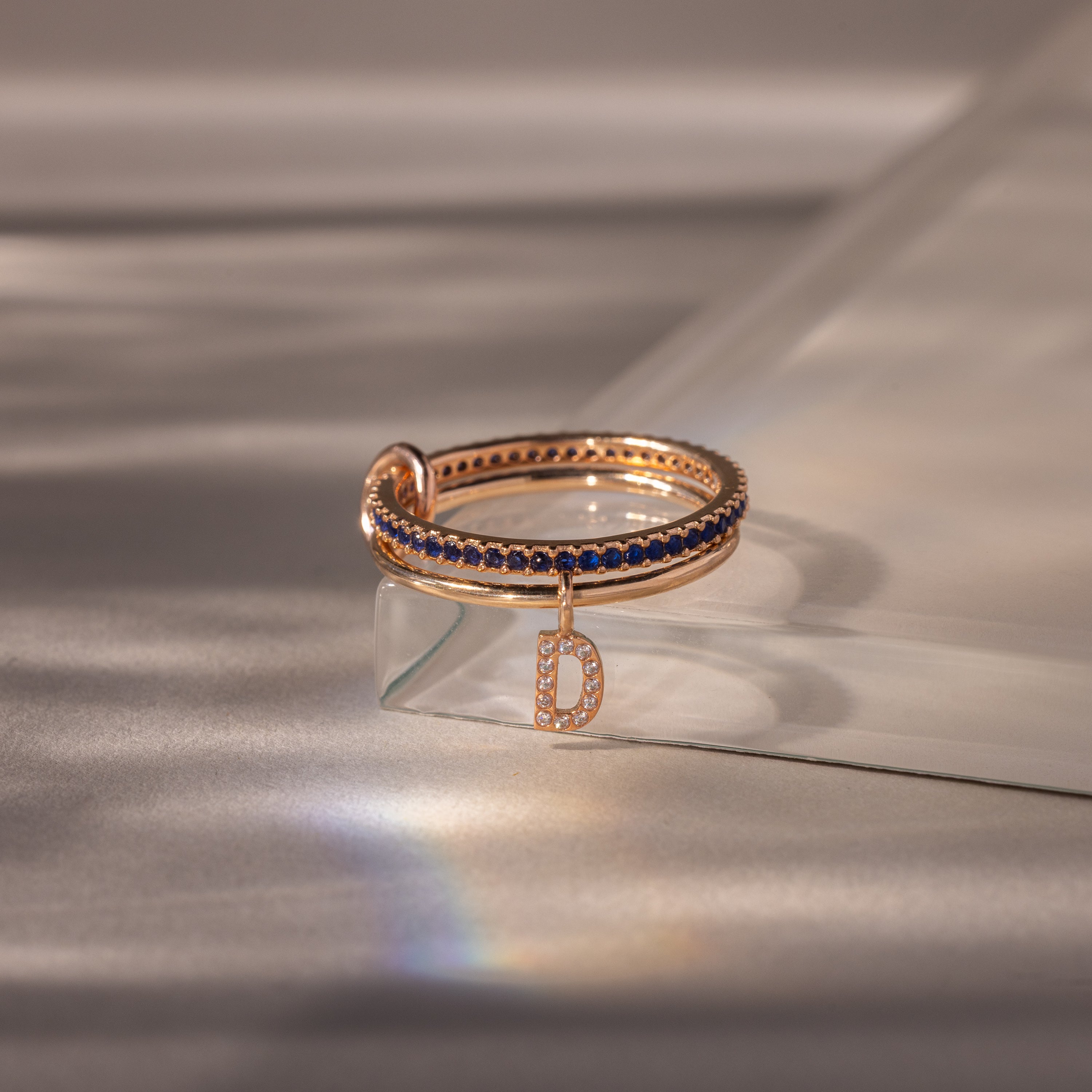 Linked Initial Sapphire Ring Set with blue stones and a diamond-studded letter D charm, displayed on a glass surface—an ideal choice for fans of personalized jewelry.