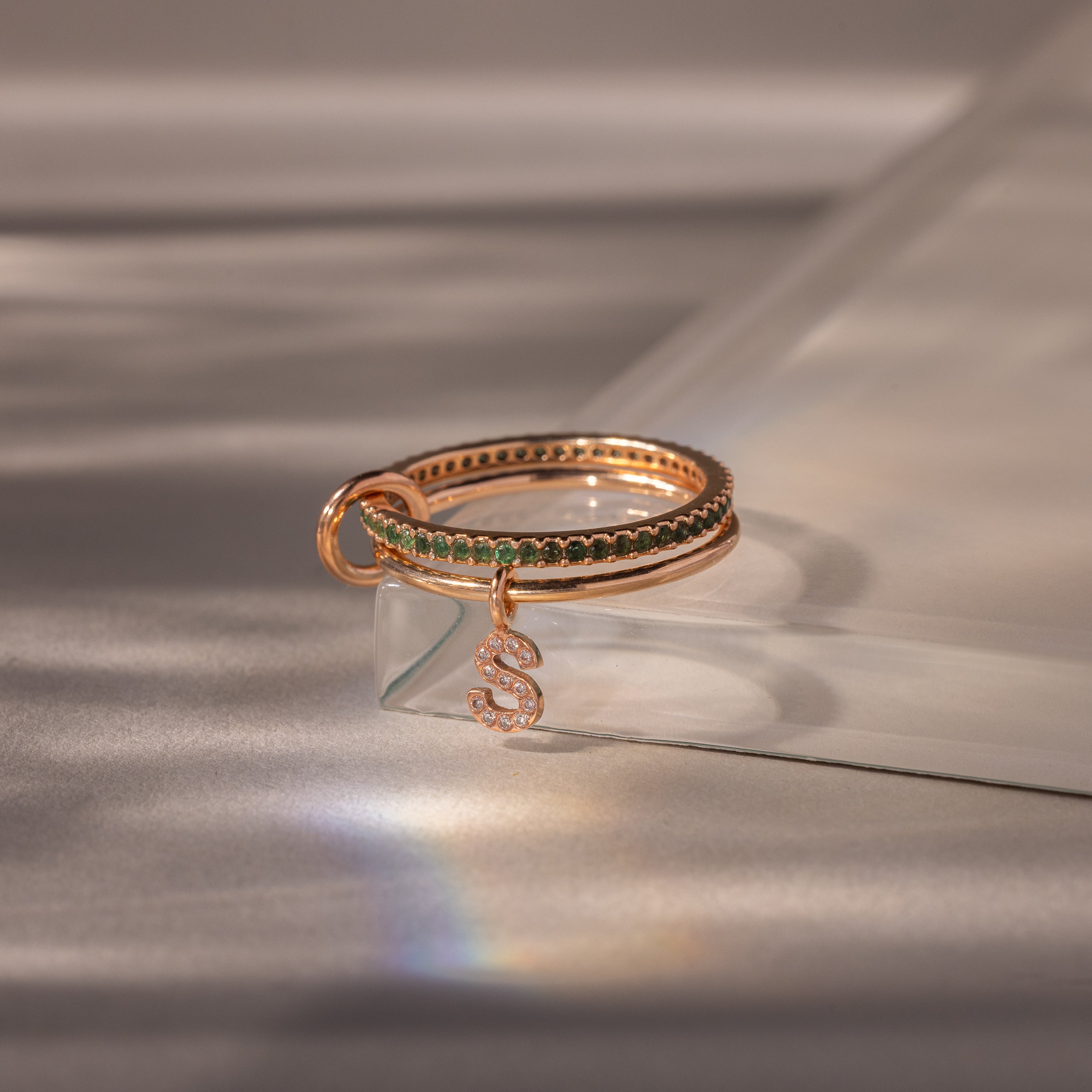 The Linked Initial Emerald Ring Set features green stones and a delicate snake charm, shown on a glass surface with soft lighting—a distinctive piece of birthstone jewelry.