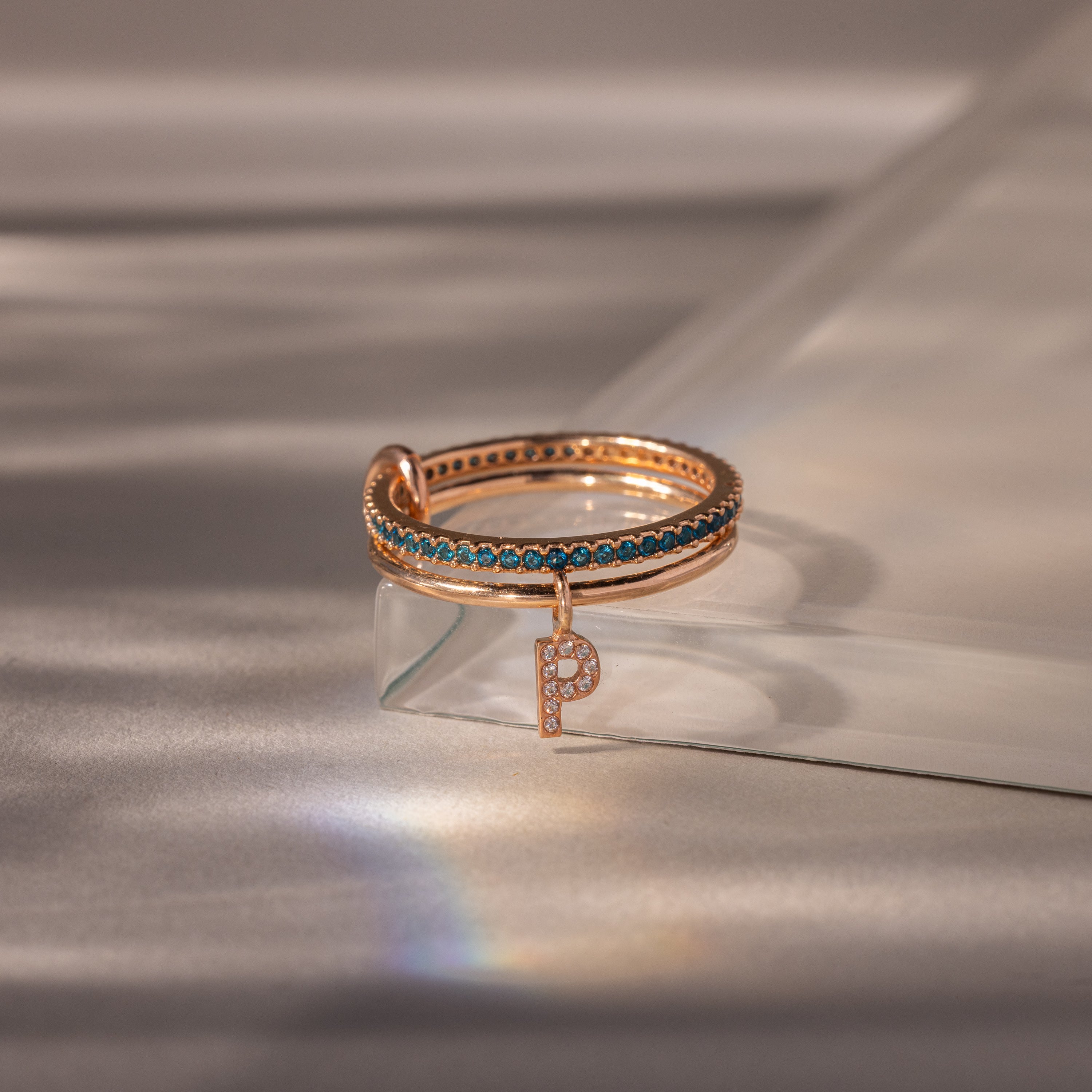 The Linked Initial Blue Zircon Ring Set showcases blue stones and a dangling, diamond-studded letter P charm on a glass surface—a stunning piece of personalized jewelry.
