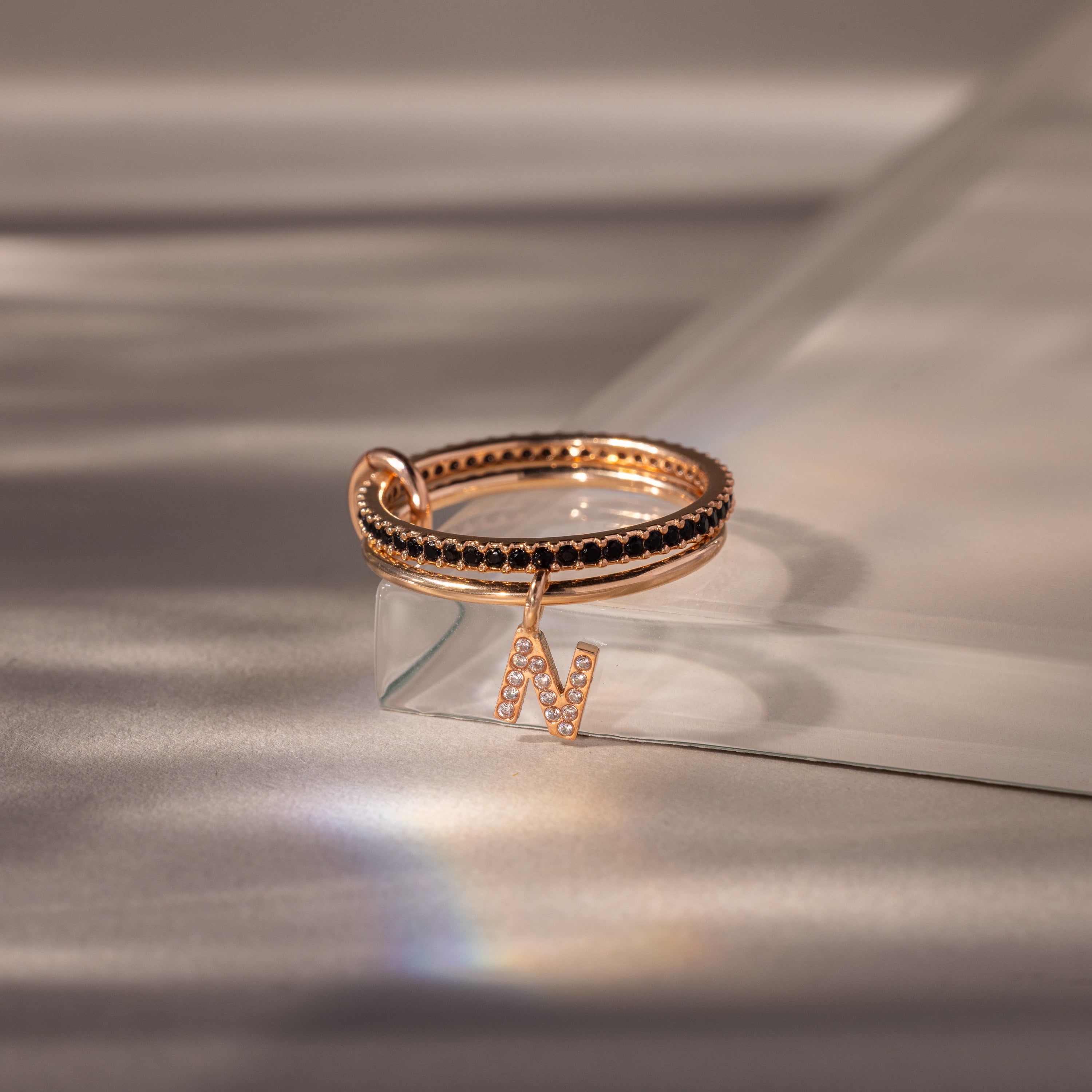 The Linked Initial Onyx Ring Set showcases a rose gold ring with a sparkling letter N charm, delicately displayed on a reflective soft surface—a beautiful piece of personalized jewelry.