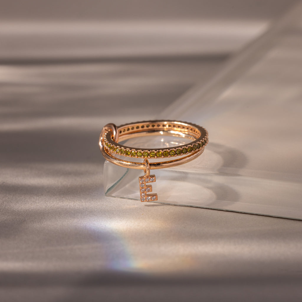 The Linked Initial Peridot Ring Set features a gold ring with a letter pendant, elegantly displayed on a clear surface—an ideal choice for personalized jewelry enthusiasts.