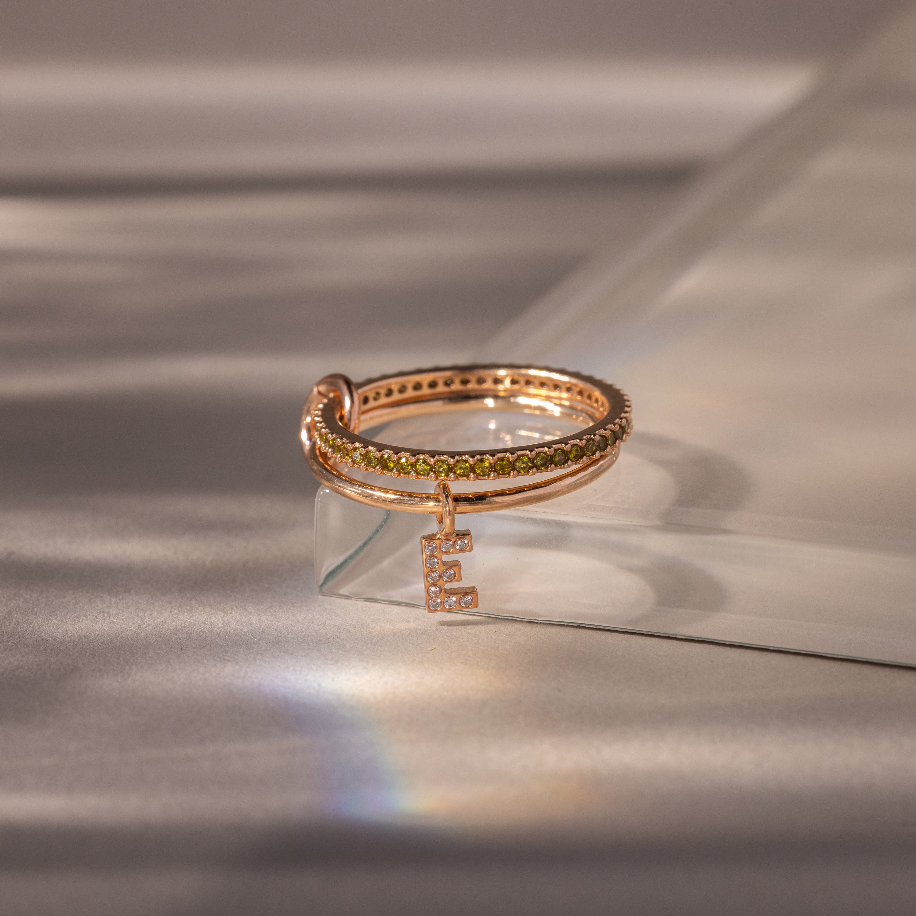 The Linked Initial Peridot Ring Set features a gold ring with a letter pendant, elegantly displayed on a clear surface—an ideal choice for personalized jewelry enthusiasts.