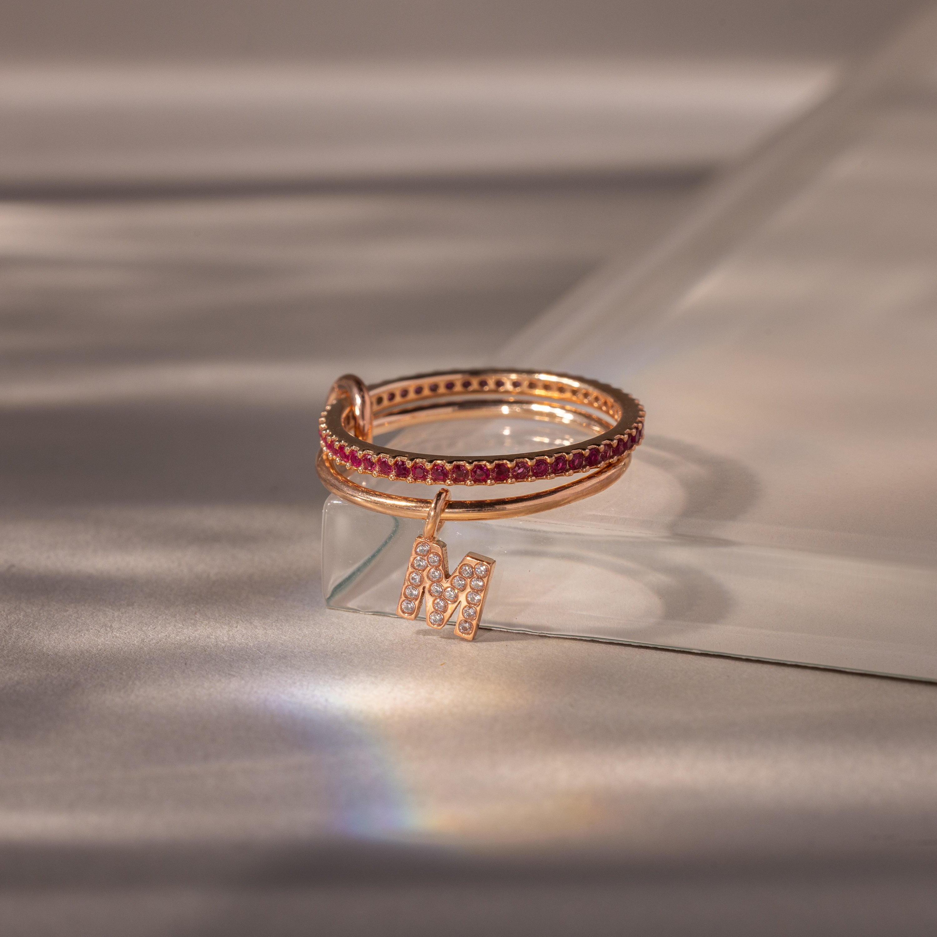 The Linked Initial Ruby Ring Set showcases vibrant red stones on a clear surface, ideal as a personalized birthstone ring or paired with a pavé initial charm for extra sparkle.