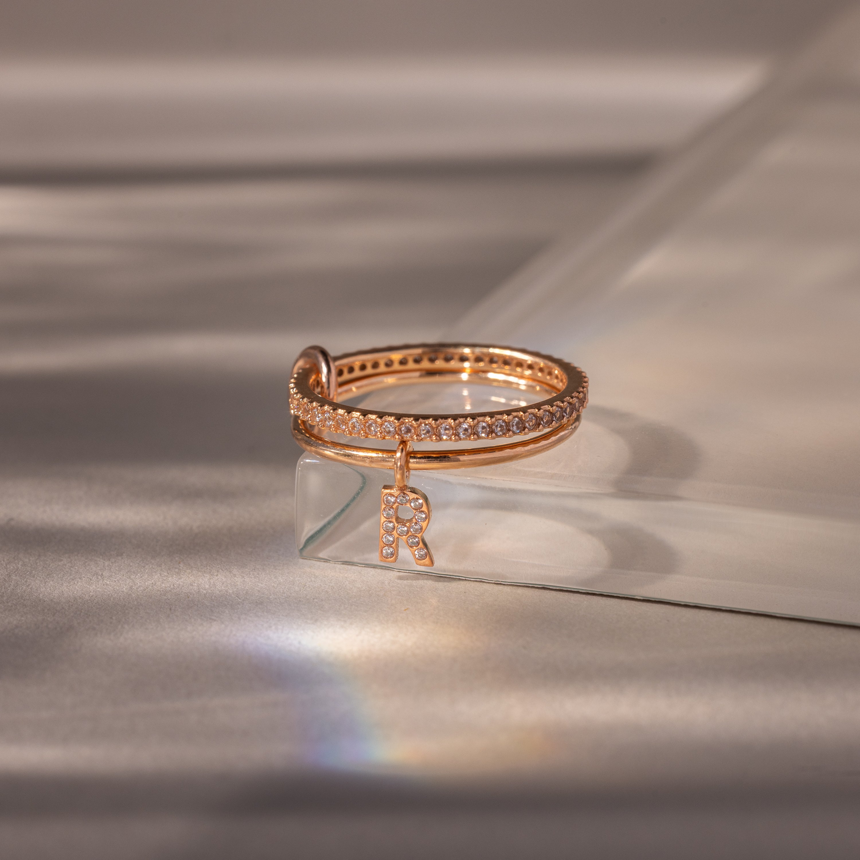 The Linked Initial Diamond Ring Set featuring small diamonds, a crystal charm, and a pavé initial "R" charm sits elegantly on a reflective surface.