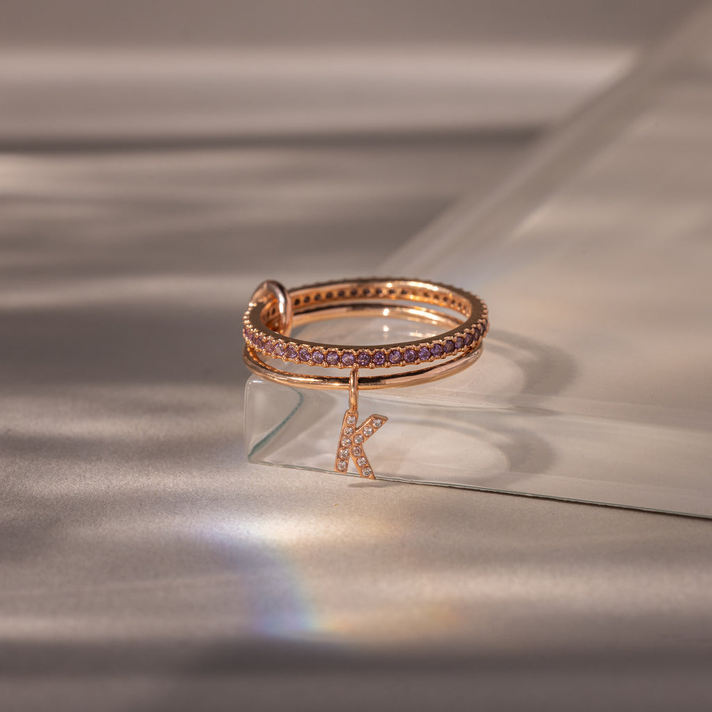 The Linked Initial Alexandrite Ring Set features rose gold bands with purple Alexandrite stones and a dangling letter K charm, elegantly displayed on a glass surface.