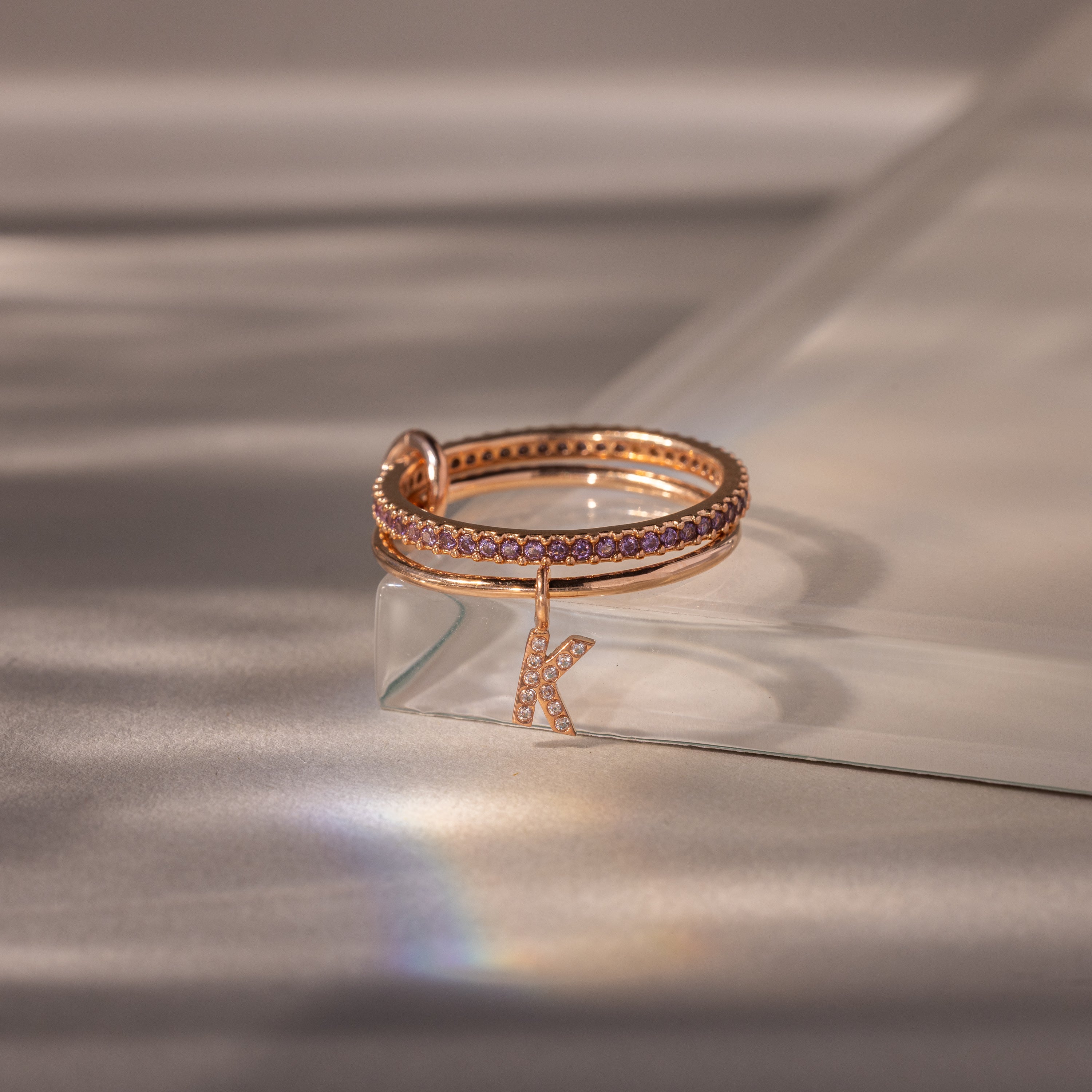 The Linked Initial Alexandrite Ring Set features rose gold bands with purple Alexandrite stones and a dangling letter K charm, elegantly displayed on a glass surface.